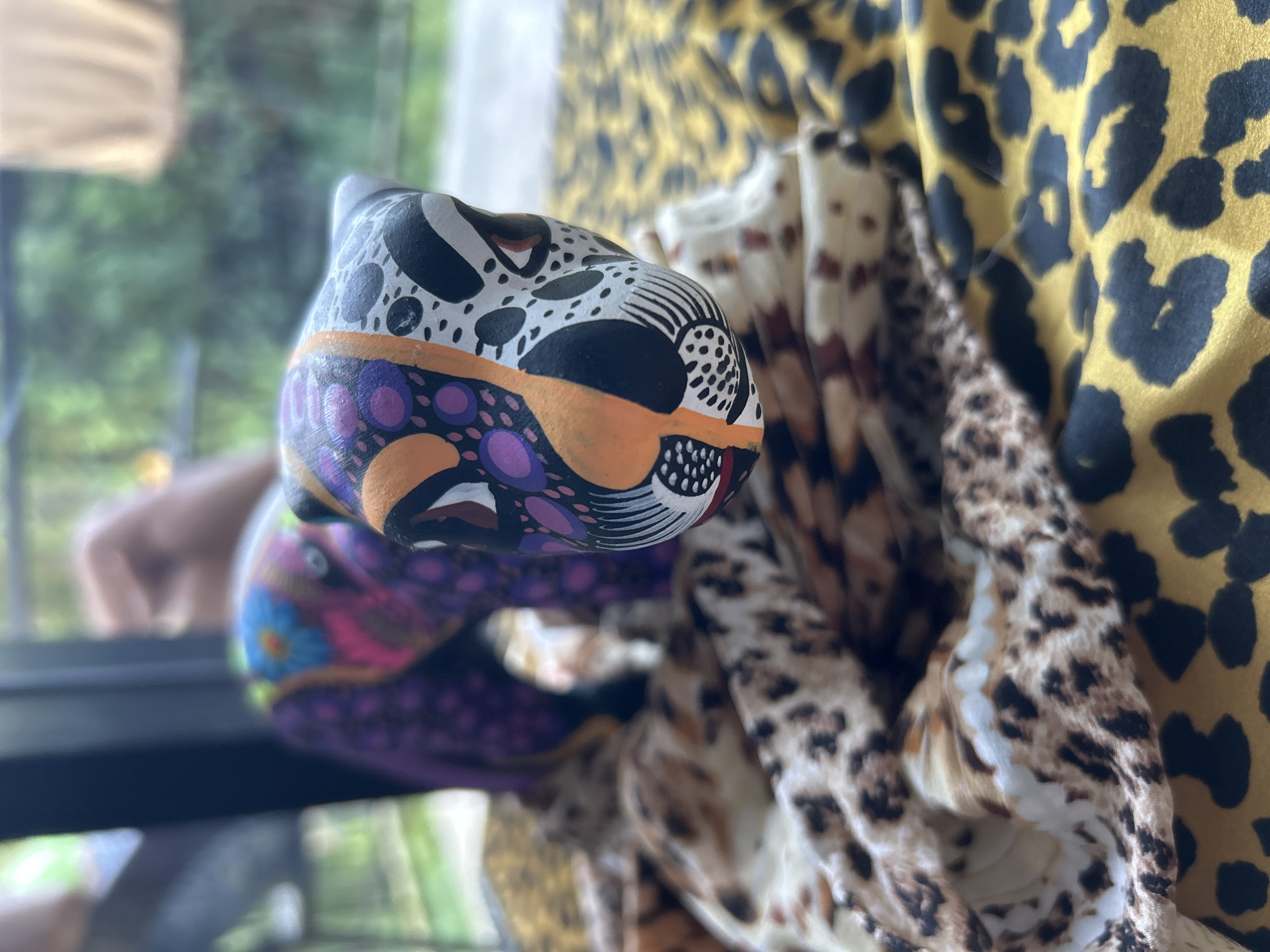 The image captures a vibrant, close-up composition featuring a colorful, stylized animal sculpture alongside draped fabrics with animal print patterns.
In the foreground, a brightly painted sculpture, possibly ceramic or wood, dominates the left side of the frame. It features intricate patterns and a palette including black, white, orange, purple, pink, and blue, with various dots, lines, and abstract shapes suggesting a feline head or body. The sculpture's vivid colors and design elements evoke a sense of traditional or folk art.
To the right, several fabrics with animal print patterns are draped. One prominent fabric is a bold yellow with distinct black leopard spots. Adjacent to it, other fabrics display more naturalistic brown and beige leopard or cheetah prints, creating a texture-rich backdrop.
The background is heavily blurred, revealing an outdoor setting with lush greenery, suggesting the image was taken near a window or an open-air space. A blurry human hand or arm is visible in the distant left background. The natural light indicates it is daytime. No specific activity is occurring; the image appears to be a still life focusing on the objects and their textures. There is no visible text.
Given the location context of Baan Wok Tum, Thailand, the colorful sculpture and natural setting are consistent with local artistry and the tropical environment.