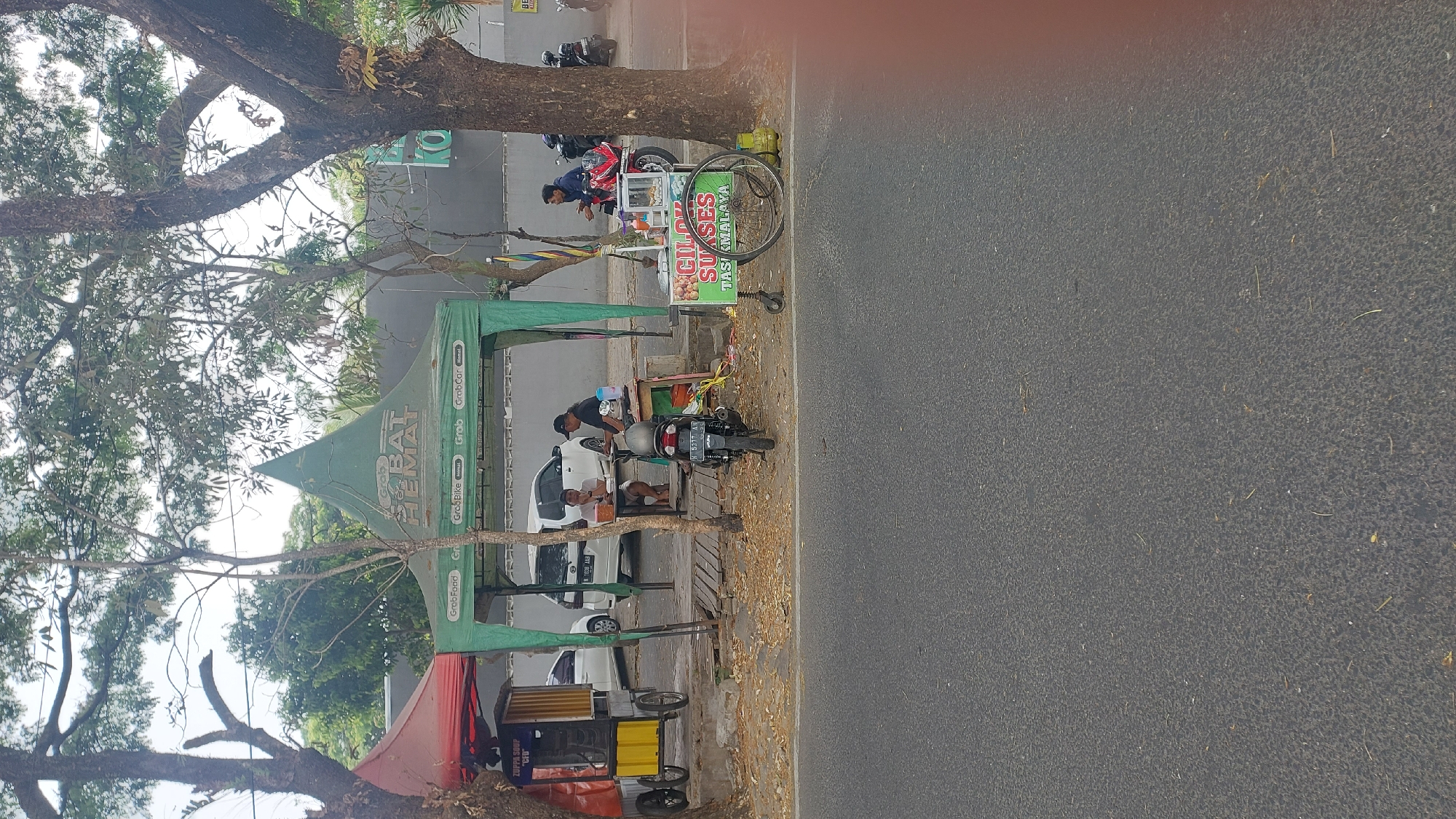 The image depicts a roadside scene in Malang, Indonesia, featuring a food stall area under a green tent with "Grab Hemat" branding. Two individuals are seated at a table under the tent, seemingly eating or conversing. A black motorcycle is parked in front of them. To the left, a red tent covers a food cart labeled "ZUPPA SOUP 'CFD'". Further to the right, another food cart with the label "CILOK SUKSES TASIKMALAYA" is visible, with its wheel turned sideways. A person in a blue jacket is seated on a red motorcycle parked near this cart. In the background, a white car is parked, and trees line the street. The overall environment appears to be a typical roadside commercial area in Malang.