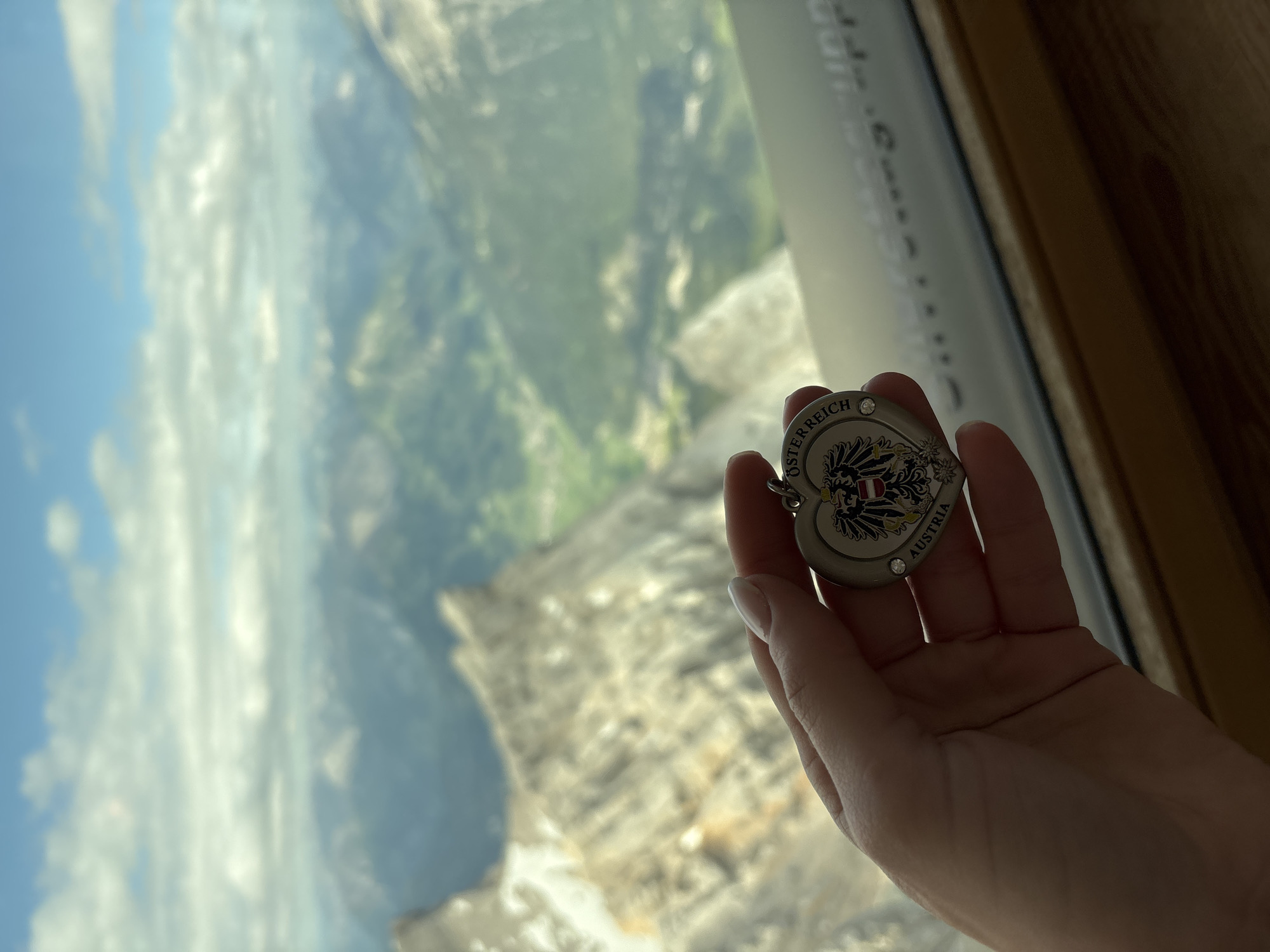 A hand is holding a heart-shaped keychain with the Austrian coat of arms and the text "ÖSTERREICH" and "AUSTRIA". The keychain is silver and has small rhinestones. The background is a panoramic view of the Alps, with blurred mountains and a cloudy sky. The scene is likely from the Zugspitze, which is the highest peak in Germany and borders Austria, and is a popular tourist destination accessible from Ehrwald, Austria.
