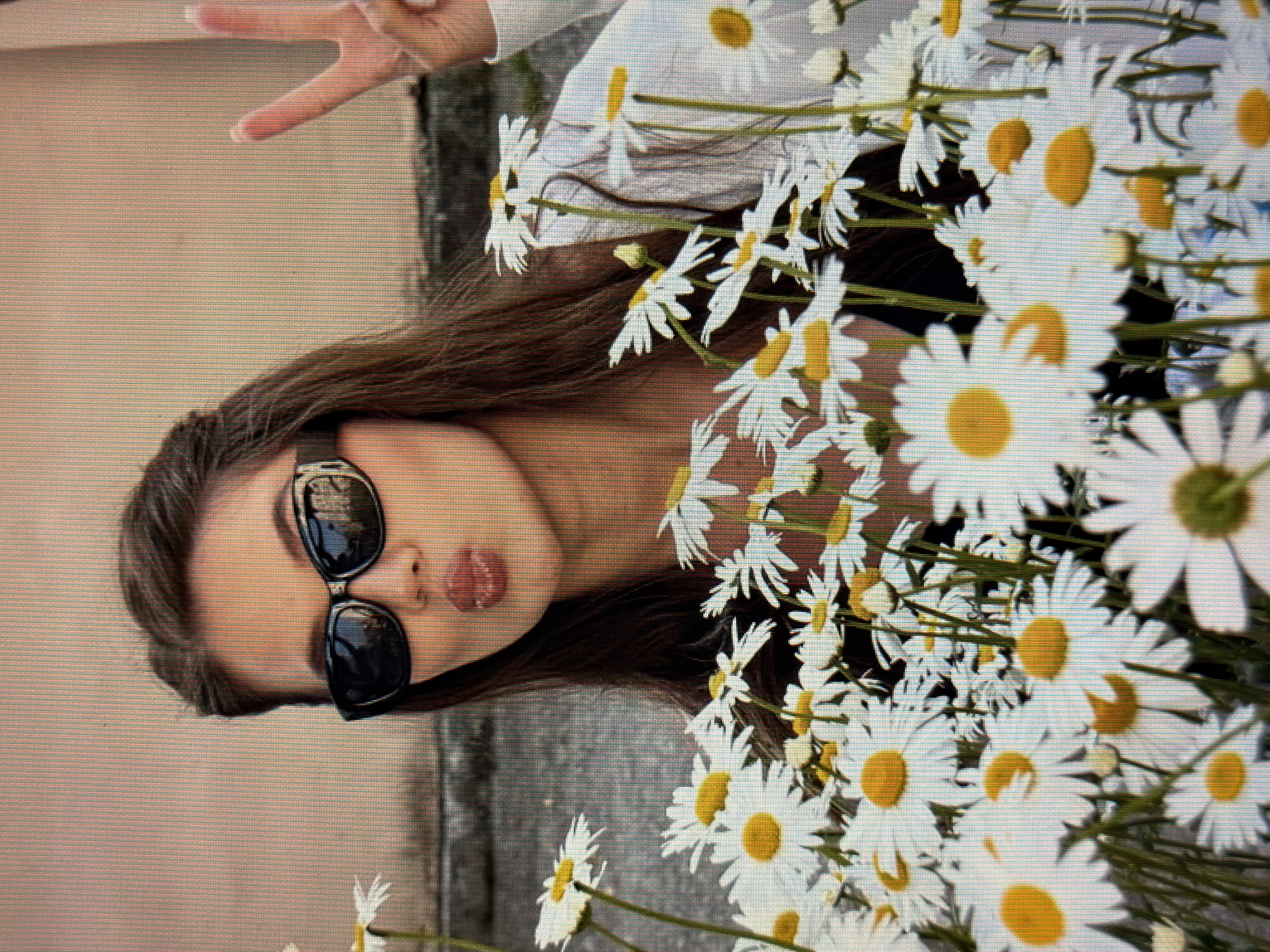 A young woman with brown hair wears black sunglasses and a white shirt, with a black top underneath. She is holding up two fingers in a peace sign with her right hand, and her lips are pursed as if blowing a kiss. She is surrounded by a large bouquet of daisies. The background appears to be a light-colored wall with a hint of a grey surface below, possibly a sidewalk or building facade. There is no information in the image to indicate a specific location in the city of Sirius, Russia.