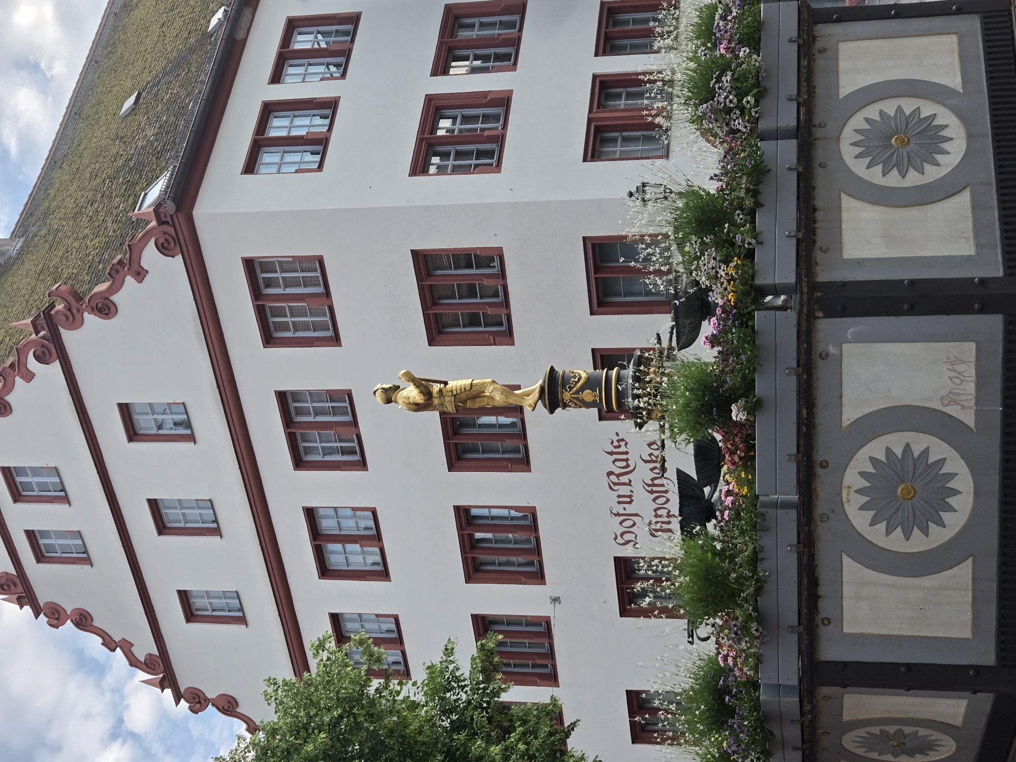 The image captures a vertical view of a historic building facade with a prominent, ornate fountain in the foreground. The building is white with deep red trim around numerous evenly spaced rectangular windows. The roof is tiled and appears to be covered in moss. Decorative red molding adorns the eaves of the building.
The fountain features a golden statue of a figure in armor, seemingly poised for action, atop a dark, decorative column. Below the statue, the building has the German text "Hof-u.-Rats-Apotheke" written in elegant, dark red lettering. Lush greenery and a profusion of colorful flowers in planters surround the fountain and cascade down its sides, suggesting a well-maintained public space.
The scene is set outdoors, likely during daylight hours. The sky is partly cloudy with patches of blue, indicating fair weather. The architectural style and the signage suggest the location is in Germany, specifically Ansbach, as indicated by the provided context. The overall impression is one of historical charm and vibrant urban life, with the fountain acting as a central focal point amidst the classic architecture. The image is rotated 90 degrees counterclockwise.