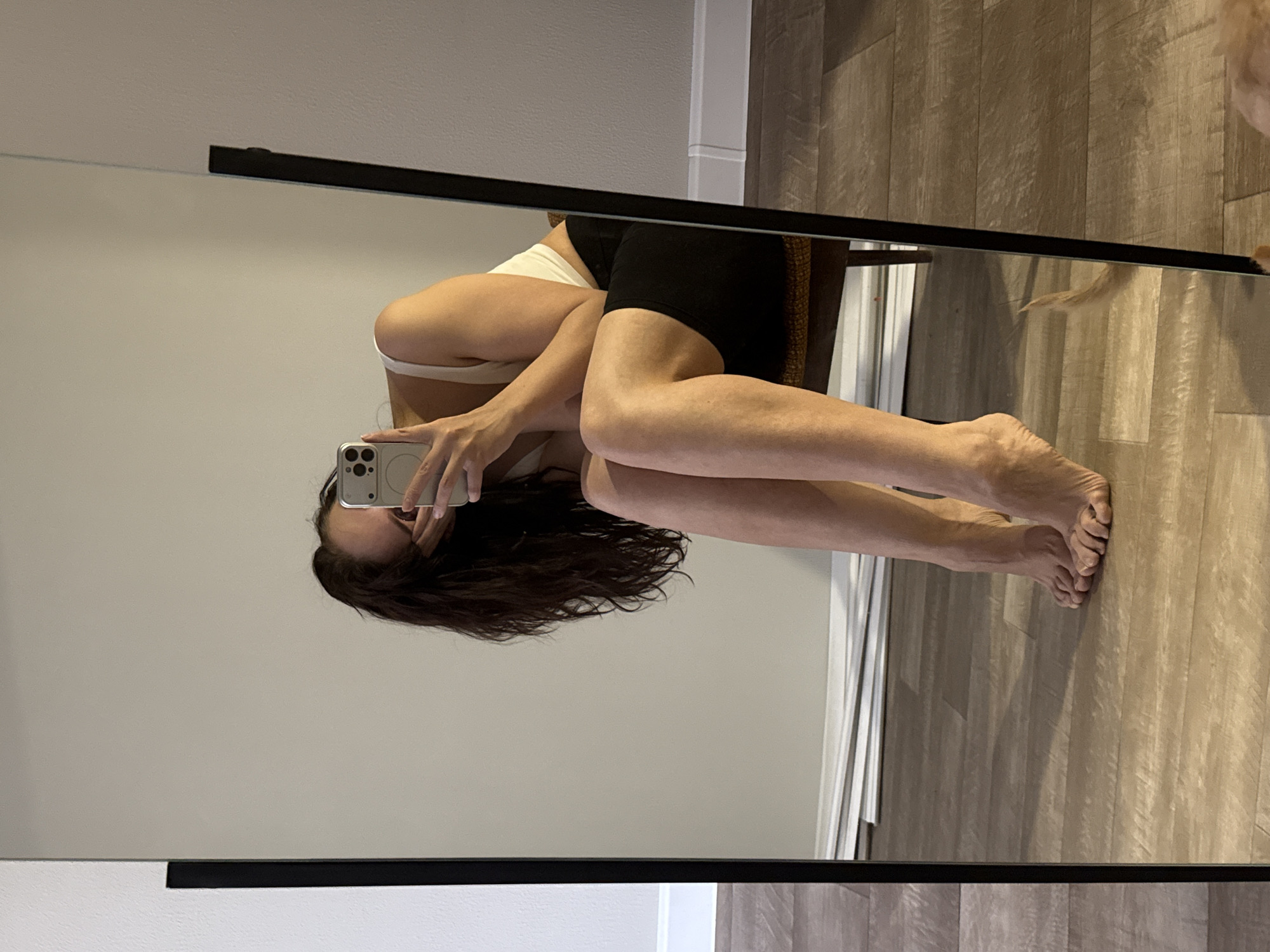 The image is a mirror selfie. A woman in a white sports bra and black shorts is sitting in a chair, facing the mirror, with her legs extended towards the right. She is holding a cell phone up in front of her face to take the picture. The background shows a gray wall and a light-colored wood floor. The location is not discernible from this image.