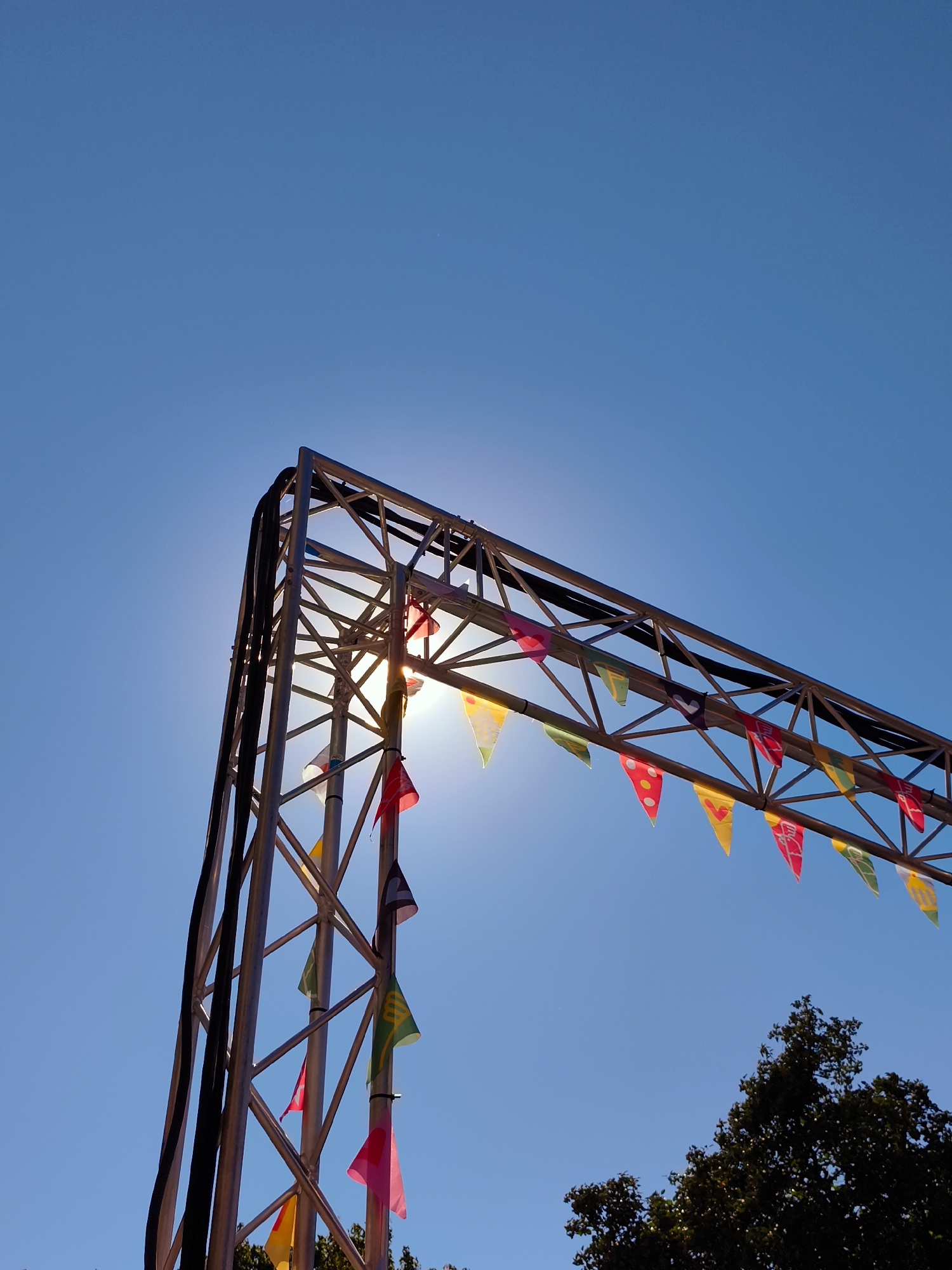 The image shows a close-up, low-angle shot of a metal truss structure against a clear, bright blue sky. The sun is partially obscured by the truss, creating a lens flare effect. A string of colorful triangular flags is draped along the top horizontal beam of the truss. The flags are a mix of solid colors and some have patterns or designs, including what appear to be stylized characters or symbols. To the lower right of the truss, the dark green foliage of a tree is visible against the sky. The overall impression is of an outdoor event or stage setup, likely in a park or open area. There are no people or any indications of a specific location in Gdańsk, Poland.