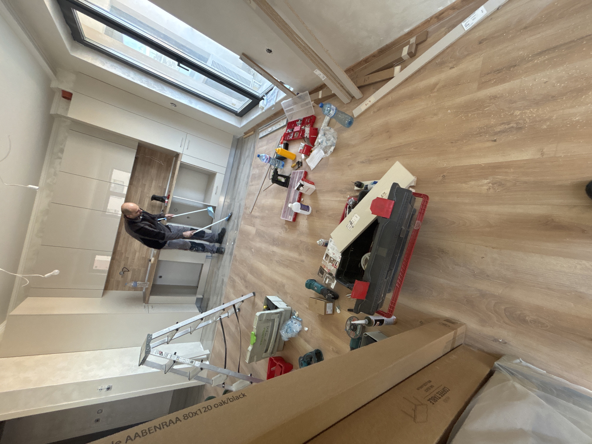 A man is installing cabinets in a room with a sloped skylight. The room has light wood flooring and unpainted walls. Construction materials, tools, and empty water bottles are scattered around the floor. A ladder is positioned near the cabinets. The bottom of boxes are visible in the foreground and at the bottom of the frame is an "AABENRAA 80x120 oak/black" label. The location is likely a home renovation site in Plovdiv, Bulgaria, but nothing confirms this location.