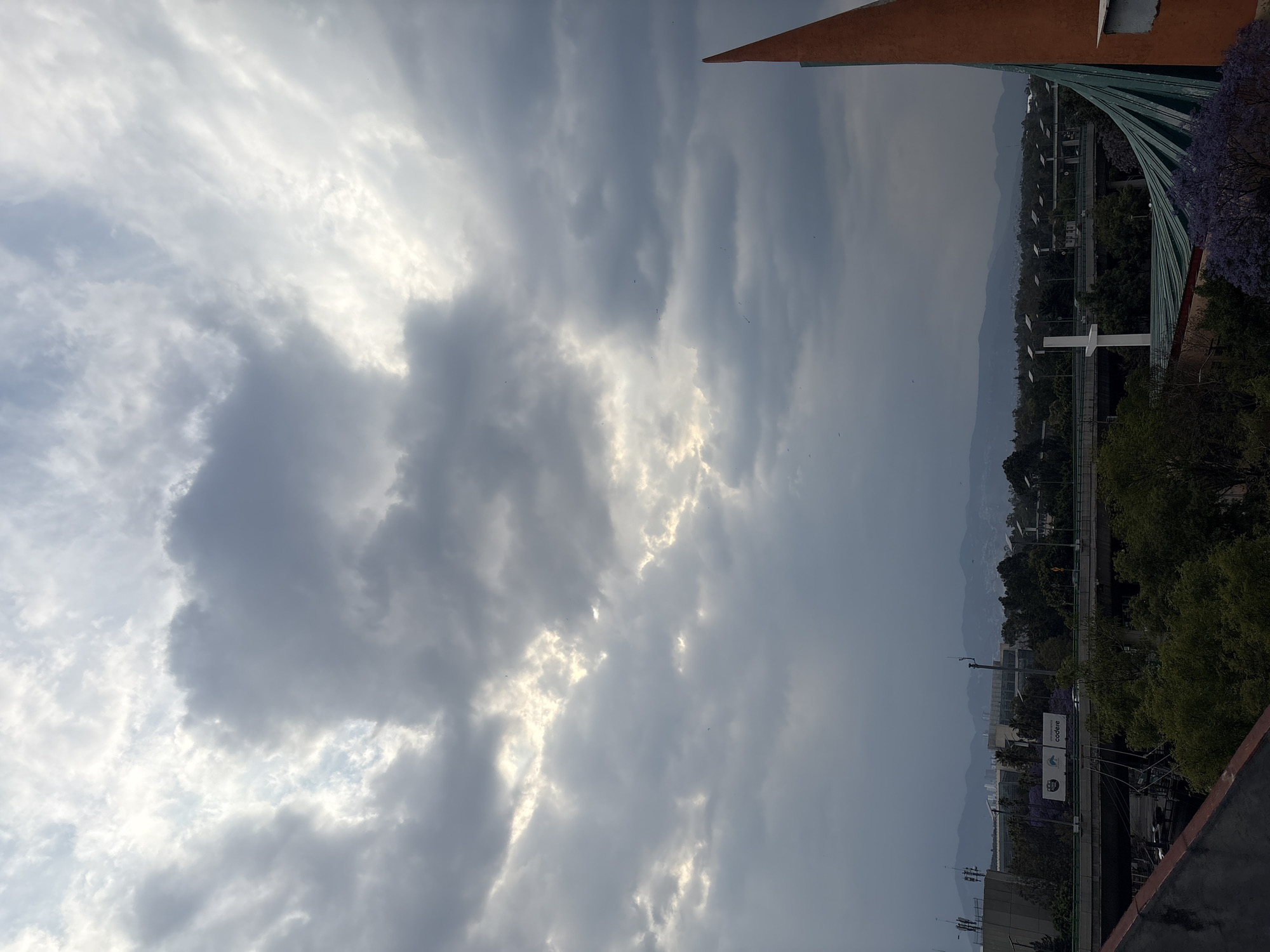 The image depicts a cloudy sky above a cityscape in Mexico City, Mexico. A highway with cars is visible, along with buildings and trees. A tall white cross stands near the highway. Parts of a structure with a green, rib-like design and a rusty-orange triangular shape are visible at the top of the frame.