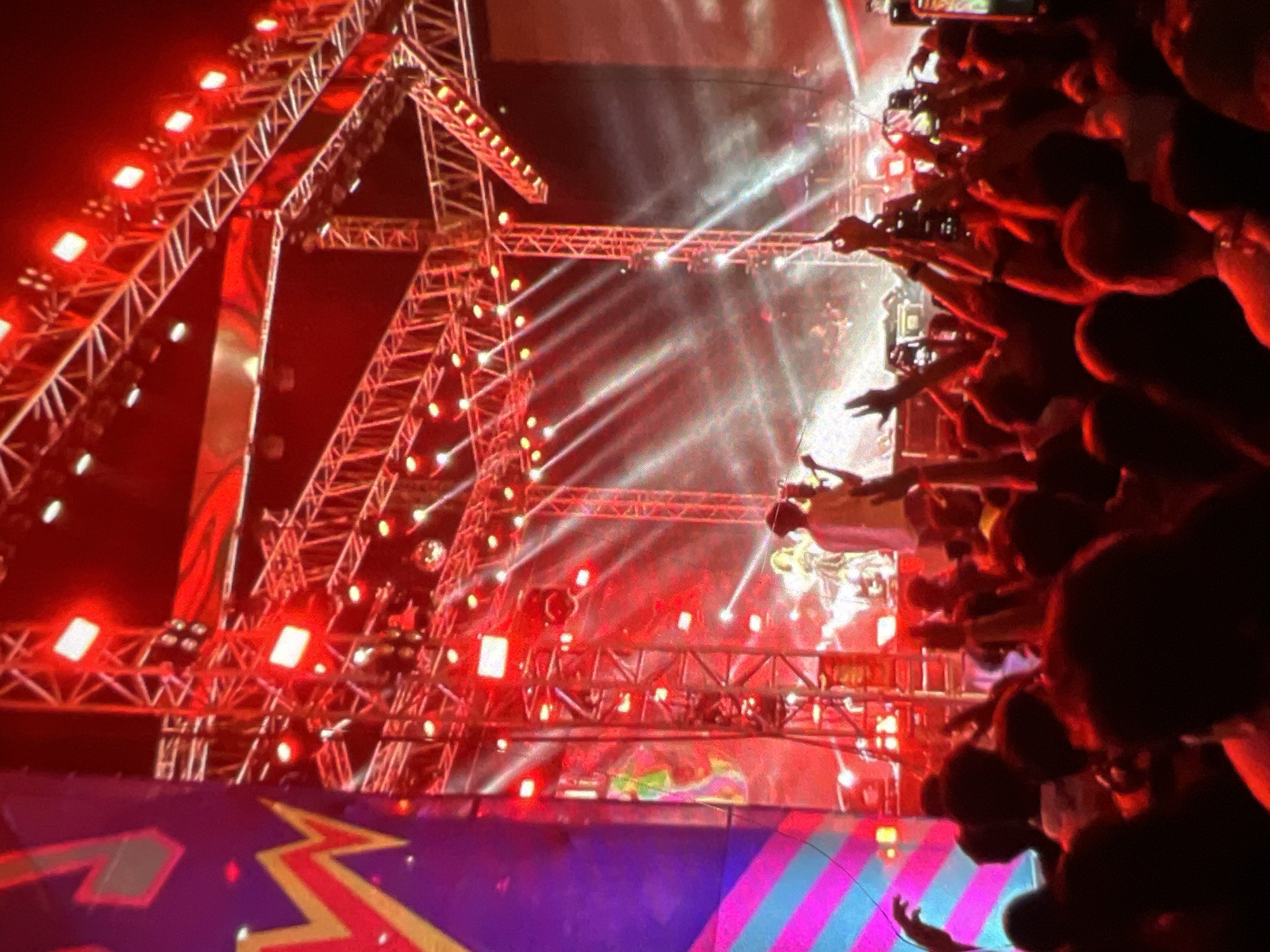 The image captures a vibrant concert scene with a large crowd gathered in front of a stage adorned with numerous bright red lights. The stage structure is complex, featuring an intricate network of metal scaffolding and lighting rigs that illuminate the venue with a dramatic red glow. Beams of white light cut through the smoky atmosphere, creating a dynamic and energetic ambiance. In the background, on the stage, two figures are visible, seemingly performing or interacting with the audience. One figure is wearing a light-colored garment, while the other is in darker clothing. To the left of the stage, a large, colorful banner or screen displays abstract geometric patterns in shades of purple, pink, and yellow. The crowd in the foreground is densely packed, with many individuals raising their hands, some holding up mobile phones to capture the event. The overall impression is one of excitement and shared experience at a live musical performance. The context of Lagos, Nigeria, suggests this is likely an outdoor or large indoor venue hosting a popular event, potentially a music festival or concert by a well-known artist.