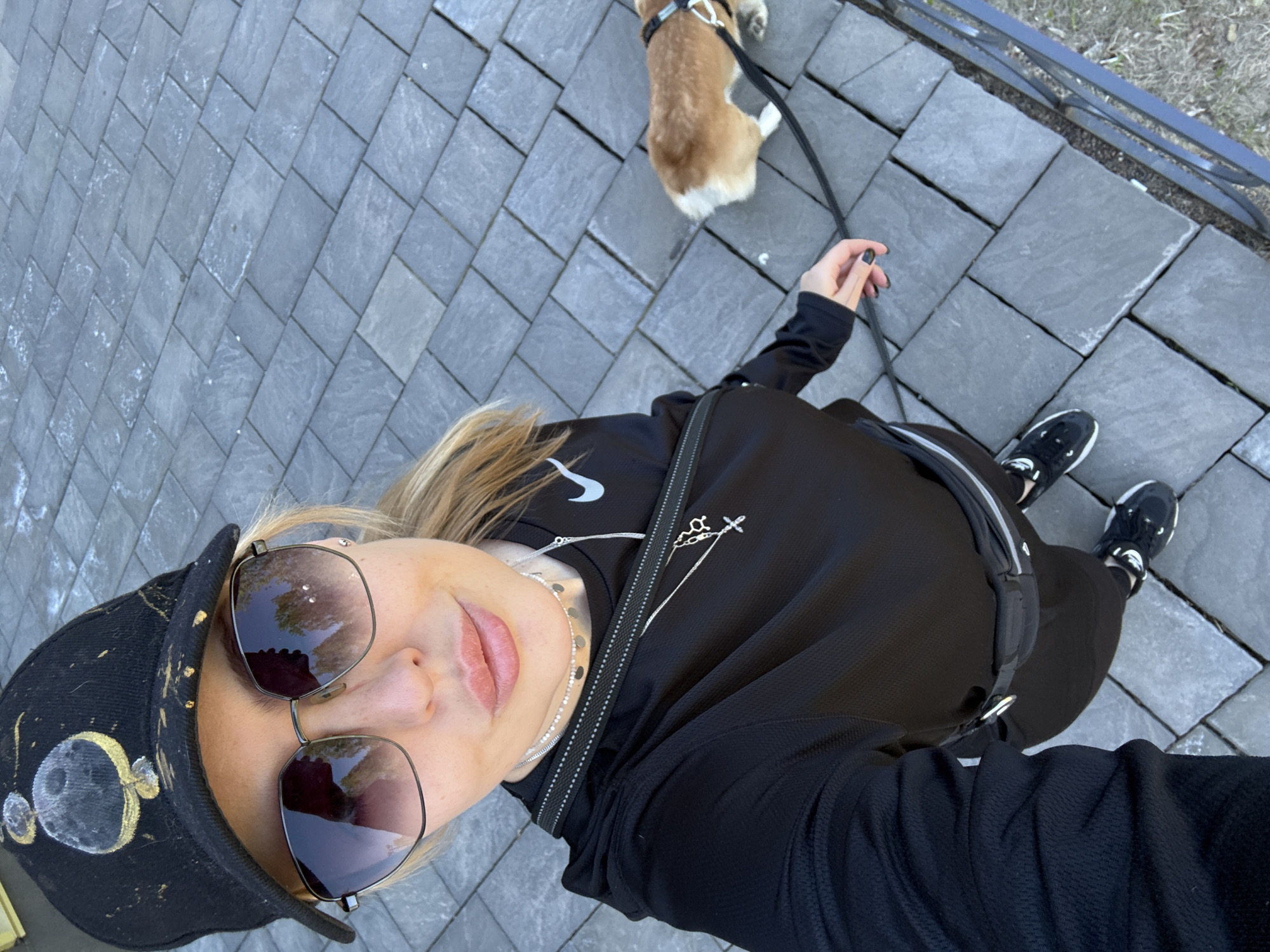 An overhead selfie captured outdoors shows a person walking a small dog on a leash on a sunny day in what appears to be Krasnoyarsk, Russia. The person, a blonde woman, is seen from the chest up, wearing large reflective sunglasses, a black baseball cap adorned with white and gold circular patterns (resembling moons), and a black long-sleeved top with a zipper. A white Nike swoosh logo is faintly visible on her left shoulder. She wears multiple silver necklaces, one featuring a small cross pendant and another with disc-like elements. Her lips are slightly parted, showing a relaxed expression. Her right hand, with black nail polish, holds the leash.
The dog, a small, light brown and white breed (possibly a Corgi), is seen from above walking ahead of the person, wearing a harness. The ground beneath them is a paved walkway composed of neatly laid gray rectangular tiles. In the upper right corner of the image, beyond the paved area, a dark metal fence runs alongside some dry grass, suggesting the edge of a park or outdoor area. The clear reflections in the sunglasses and the overall bright lighting indicate daytime and pleasant weather. The image is taken from a high angle, looking down at both the person and the dog.