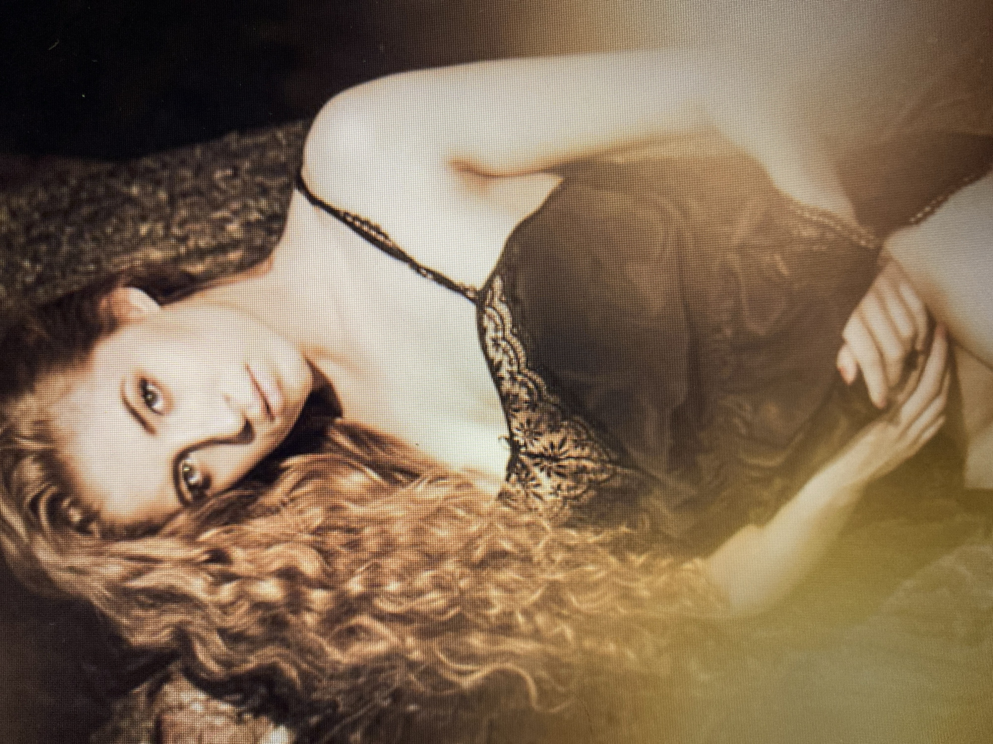 A woman with long, curly, auburn hair is positioned outdoors, leaning against a tree. She is wearing a black lace-trimmed camisole and has her hands clasped in front of her. The lighting is warm and the background is blurred, suggesting a shallow depth of field. The scene appears to be a portrait or artistic photograph.