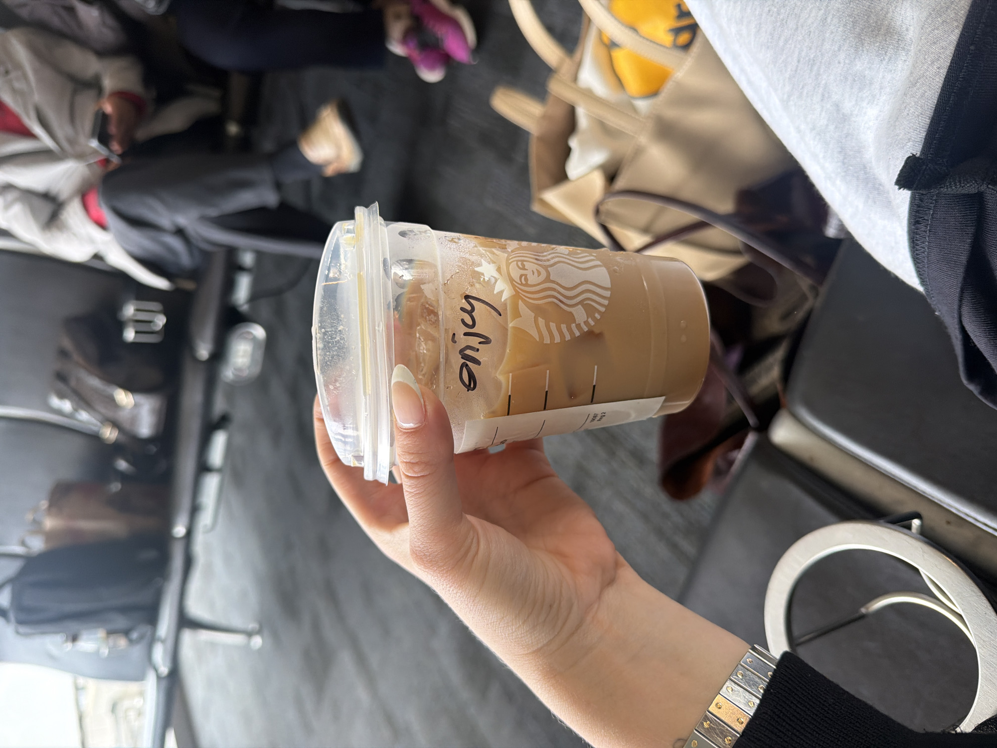 The image, captured in Los Angeles, United States, focuses on a person's hand holding a Starbucks iced coffee cup in an indoor public setting. The main subject is a clear plastic Starbucks cup, partially filled with a light brown beverage and ice. The iconic Starbucks siren logo is visible on the cup, and the word "Enjoy" is handwritten in black marker on its side. The hand holding the cup has manicured nails with French tips. A two-toned metal watch with alternating silver and gold links is worn on the wrist, which is partially covered by a black sleeve.
In the blurred background, other individuals are seated, and various personal items are visible, suggesting a waiting area such as an airport terminal or a lounge. One person in the background is wearing distinctive purple athletic shoes. A light-colored tote bag with yellow accents is also seen. The flooring appears dark, possibly carpeted. To the right of the person's hand, a silver-rimmed circular object, resembling sunglasses, is present. The overall activity involves a person holding a drink while waiting in a public space. The time of day is not discernible from the indoor lighting.