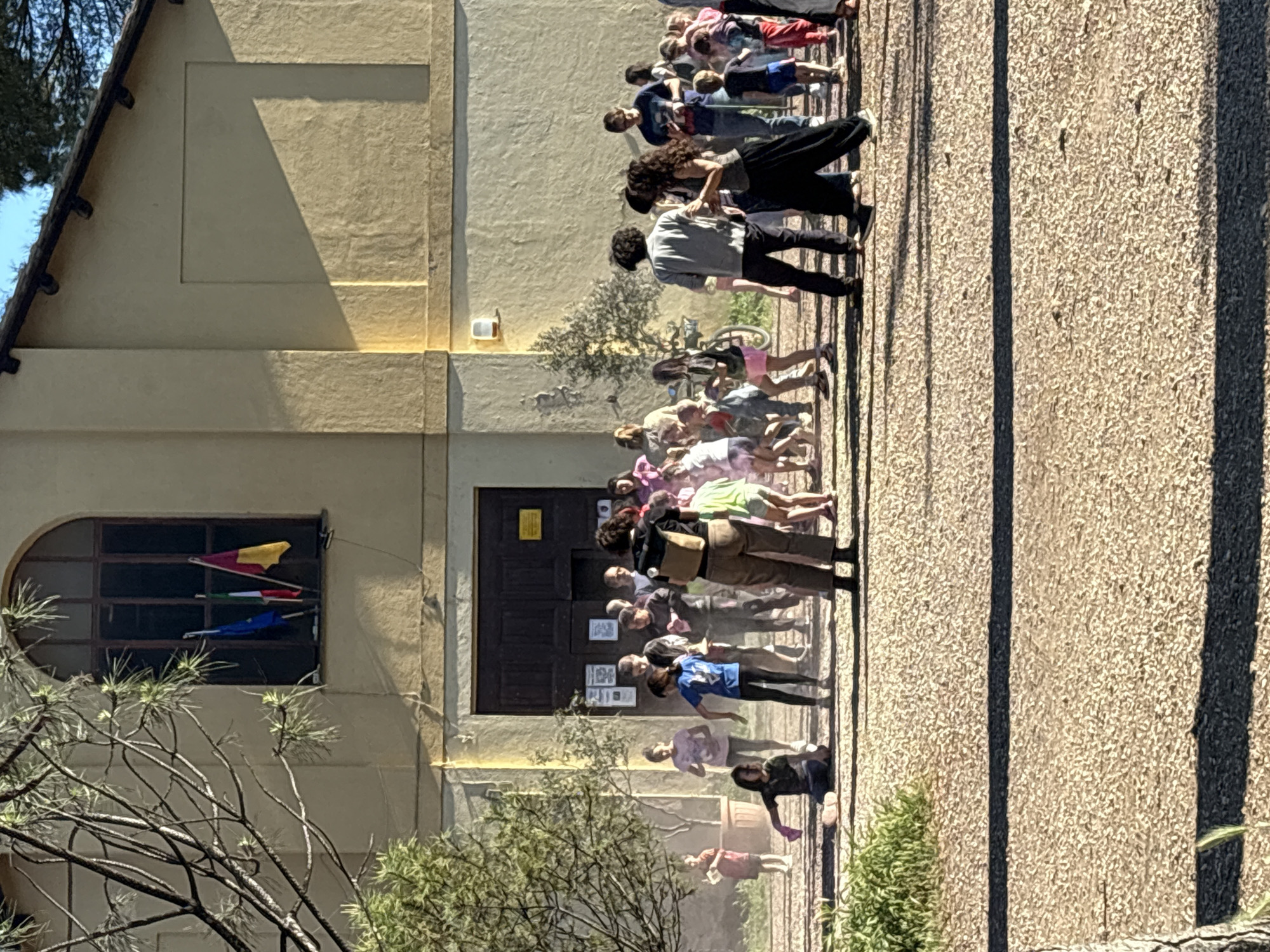 A street scene depicts a group of people standing in front of a building with a window displaying colorful banners, and a dark-colored double door entrance. The exterior of the building is a light tan color. The people are standing on an exterior surface with elongated shadows. A sparse tree is visible in the foreground to the left and another is centered in the background. The image is strongly lit from the left. The location is Rome, Italy.