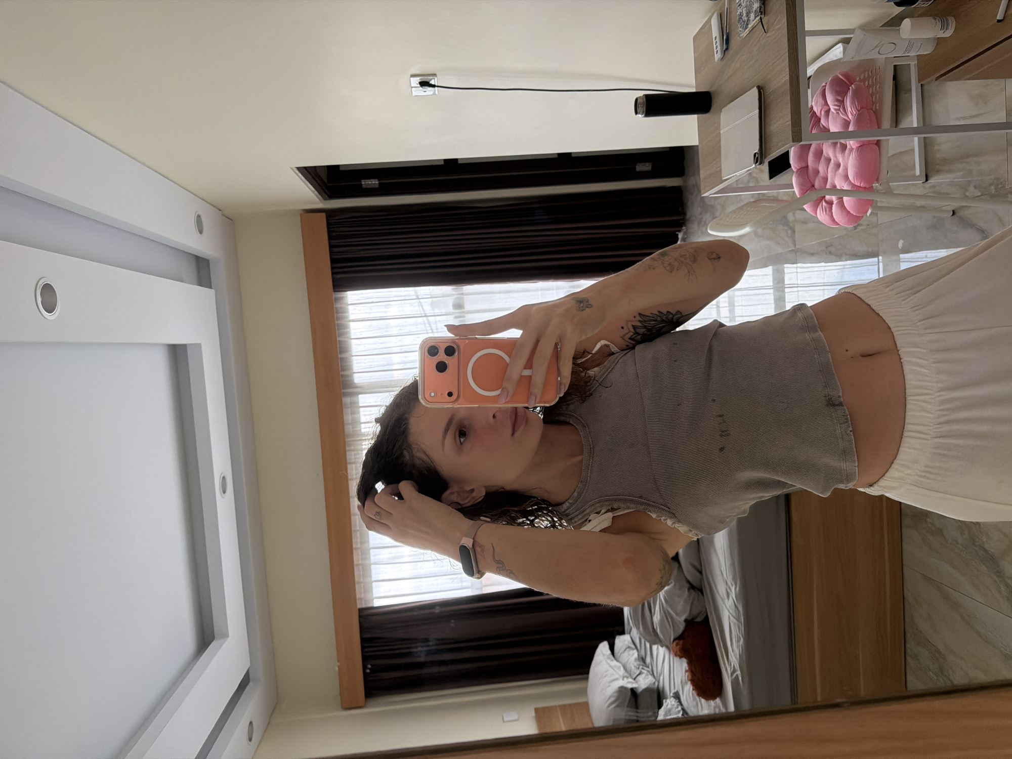 A young woman is taking a mirror selfie in a bedroom. She is wearing a grey tank top and white sweatpants. She has dark, curly hair and tattoos on her arms. She is holding an orange cell phone in her right hand and touching her hair with her left hand. In the background, there is a bed, a window with curtains, and a desk with a laptop and a pink cushion. The room appears to be well-lit and modern. The location is North Kuta, Bali, Indonesia.
