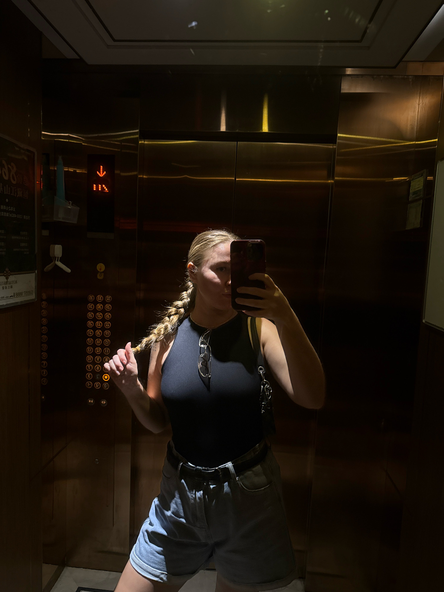 A young woman with blonde hair in a braid is taking a selfie in an elevator. She is wearing a black tank top, light blue denim shorts, and a black belt. She is holding a dark red smartphone in her right hand and her left hand is touching her braid. The elevator has wood-paneled walls and a stainless steel door. An illuminated panel with floor numbers and directional arrows is visible on the elevator wall. The lighting in the elevator is dim, with yellow light reflecting off the walls. There are signs on the left side of the elevator wall, one of which appears to be in Chinese characters. The elevator is likely located in a hotel or commercial building in Yiwu, China, based on the presence of Chinese text.