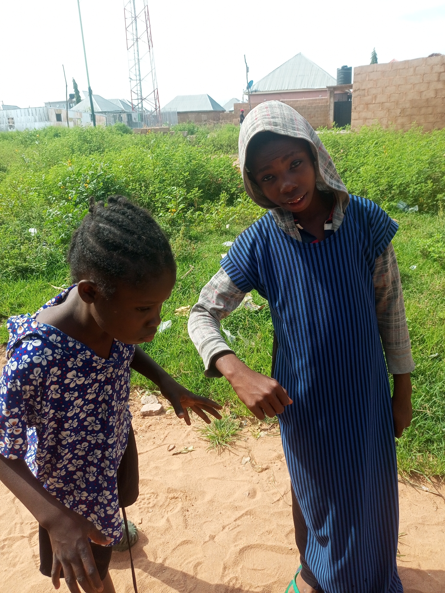 Two young children, one wearing a blue and white floral dress and the other wearing a hooded blue and black striped garment, stand on a sandy patch of ground in a semi-urban area of Tempure, Nigeria. The child in the floral dress has her hair in braids and is looking down with her hands extended. The child in the striped garment is wearing a hood and looking at the camera with a slight smile. Behind the children, a patch of tall green grass and brush is visible, with a few scattered pieces of white debris. In the background, there are various structures, including a large cell tower, several buildings under construction or in disrepair, and a brick wall. The overall impression is a sunny, outdoor environment.