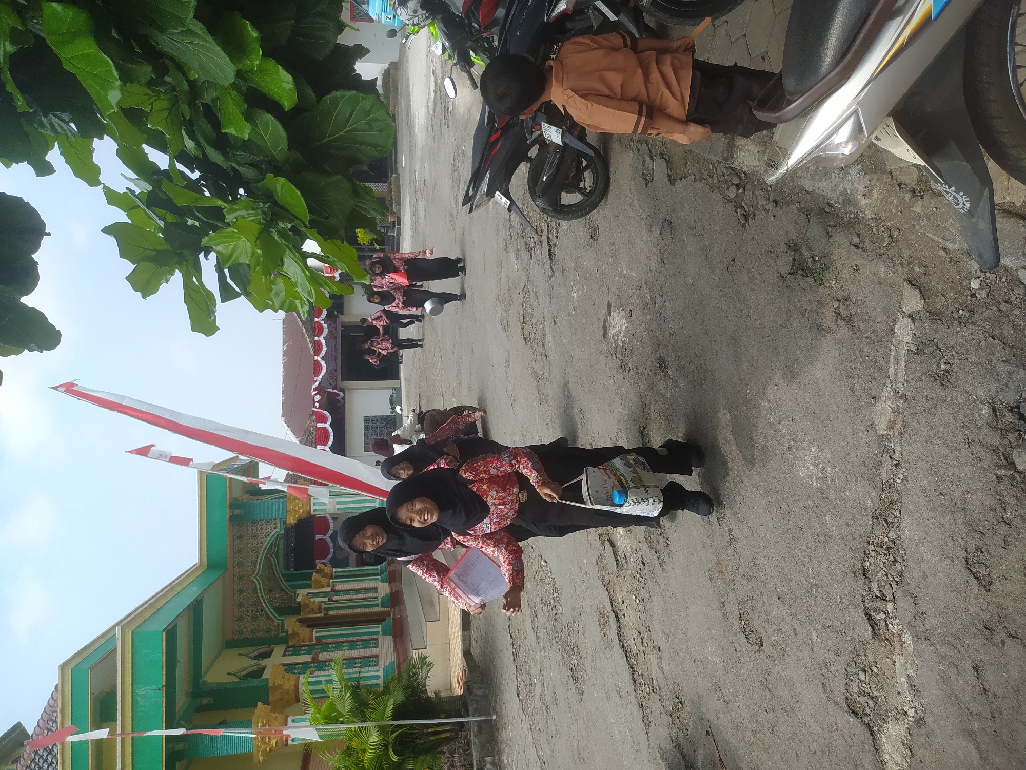 The image depicts a street scene in Candirejo, Indonesia, with several young girls in school uniforms walking on a paved road. The girls are wearing black headscarves and red and white patterned shirts with black trousers. One girl in the foreground is smiling and carrying a white bag with colorful writing and a blue water bottle inside. Another girl beside her holds some papers. In the background, other girls in similar uniforms are also visible, some walking and others on a motorcycle. To the left of the scene is a building with green and gold accents, featuring columns and an arched entrance, with Indonesian flags flying. To the right, there are several motorcycles parked, and a young boy wearing an orange shirt and dark trousers is standing and looking towards the motorcycles. The road surface is rough and cracked, with some debris. Lush green foliage from a large tree is prominent in the upper right portion of the image. The sky is overcast with some visible clouds.