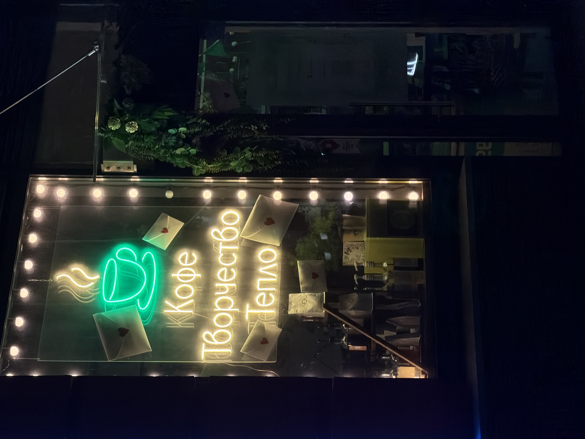 The image depicts an illuminated sign in Murino, Russia, likely an advertisement for a café. The sign, featuring a green neon coffee cup with steam and yellow neon lettering, is mounted in a window. The sign's text is in Russian and includes the words "Кофе," "Творчество," and "Тепло" – meaning Coffee, Creativity, and Warmth. There are also white envelopes with red hearts scattered around the sign. The sign is lit by a string of lights around its perimeter. Above the sign is a floral arrangement, and the building's architecture is dark and urban. The overall scene is dim, with the sign serving as the primary light source. The window and building appear to be dark at night.