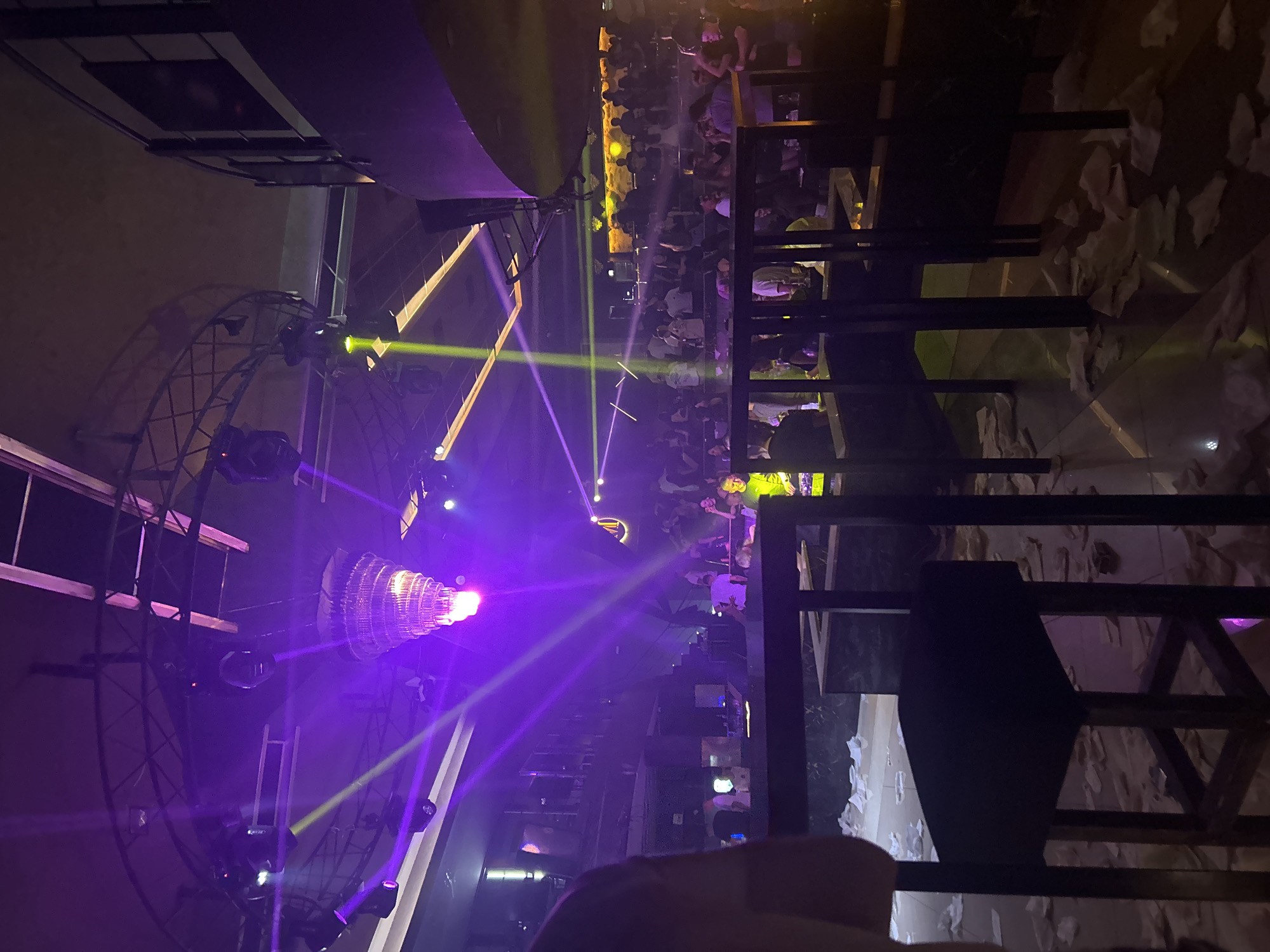 The image shows an interior scene, likely a nightclub or entertainment venue in Zlatograd, Bulgaria. The room is dimly lit with bright purple and green lighting effects. A large chandelier-like structure hangs in the center of the room, surrounded by a circular metal framework with spotlights. There are tables and chairs arranged on what appears to be an upper level of the venue. People are visible, standing and sitting in the background, suggesting a crowded event. Debris, possibly napkins or paper, lit by the club lights is on the floor near metal furniture. The perspective of the photo is from a low angle, looking up towards the stage and upper seating area. The image is rotated slightly, suggesting it was not taken upright.