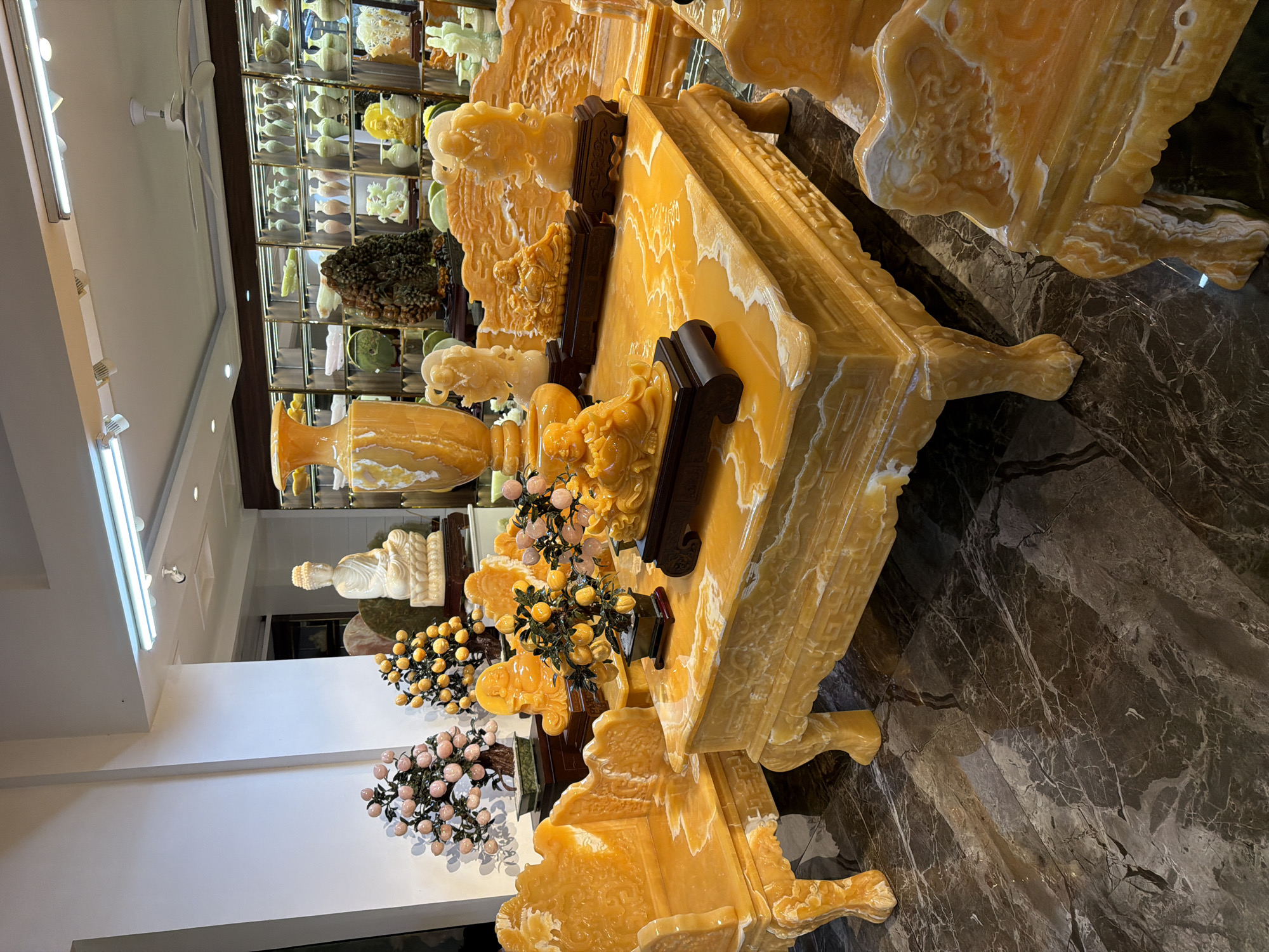 This image depicts an opulent display of yellow onyx furniture and decorative items within a well-lit showroom, likely located in Đà Nẵng, Vietnam. The focal point is a large, ornate coffee table and accompanying armchairs, all carved from translucent yellow onyx with white veining. The furniture exhibits intricate detailing, including carved legs and decorative patterns on the backrests.
On the coffee table, several decorative pieces are arranged. These include a Buddha statue on a dark wooden base, a small tree with yellow fruit, and other small carved figures also appearing to be made of onyx. Behind the coffee table, a tall, large onyx vase stands prominently. To the left of the coffee table, a seated Buddha statue carved from white stone is positioned on a pedestal. Further to the left, two decorative trees with pink and yellow fruits, also appearing to be made of stone or resin, add color to the scene.
The showroom's background features shelves stocked with numerous vases and decorative items, many appearing to be ceramics or similar materials, in various colors and sizes. Overhead lighting illuminates the space, reflecting off the polished surfaces of the onyx. The floor is a dark, polished marble with a marbled pattern. The overall impression is one of luxury and artistry, showcasing high-end decorative stone carvings. There are no people visible in the image.