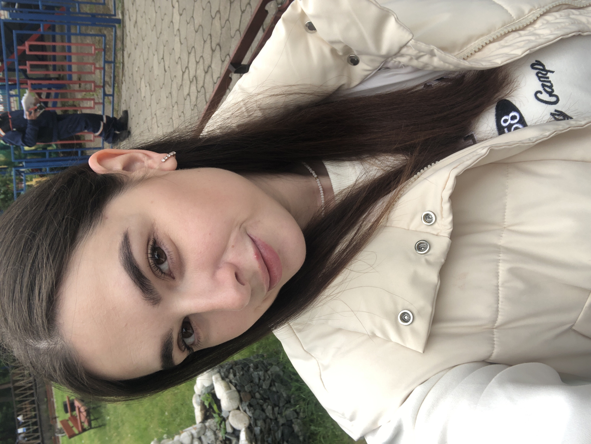 A young woman poses for a selfie outdoors in Chaikovsky, Russia. She has fair skin, dark eyebrows, and dark brown hair. She is wearing a light beige puffer vest over a white shirt, partially visible beneath the vest. The shirt appears to have a logo with the words "Camp" on it, as well as the number "58". The woman has a stud earring in her ear.
Behind her is a patch of grass and some decorative rocks and greenery. Further in the background, a multi-colored blue, red and yellow fence can be seen. A person in dark clothing is partially visible in the background, leaning against the fence. The ground appears to be paved near the fence.