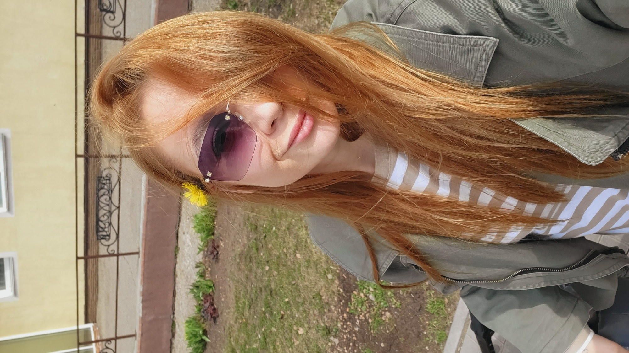 A young woman with long, red hair and sunglasses is the central figure in this photograph. She is wearing a striped top and a green jacket. A yellow flower is tucked into the side of her sunglasses. The background shows a wrought-iron fence and a building with yellow walls. There is also greenery visible, suggesting an outdoor setting. The photo was taken in the city of Chaikovsky, Russia.