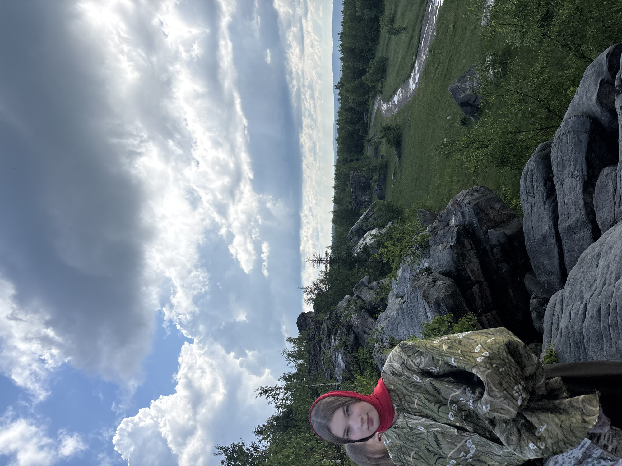 A young woman with brown hair and a red headscarf is pictured in the foreground, looking towards the camera. She is wearing a green patterned jacket. Behind her, to the left, another person is partially visible. They are standing on a rocky outcrop, with large gray rocks and green foliage surrounding them. In the background, a winding dirt road with puddles of water cuts through a grassy landscape dotted with trees. A dense forest stretches to the horizon, under a dramatic sky filled with both bright white and dark gray clouds. This location appears to be the "Ural Mountains" or "Konzhakovsky Kamen" in Gubakha, Russia, known for its rocky terrain and scenic views.