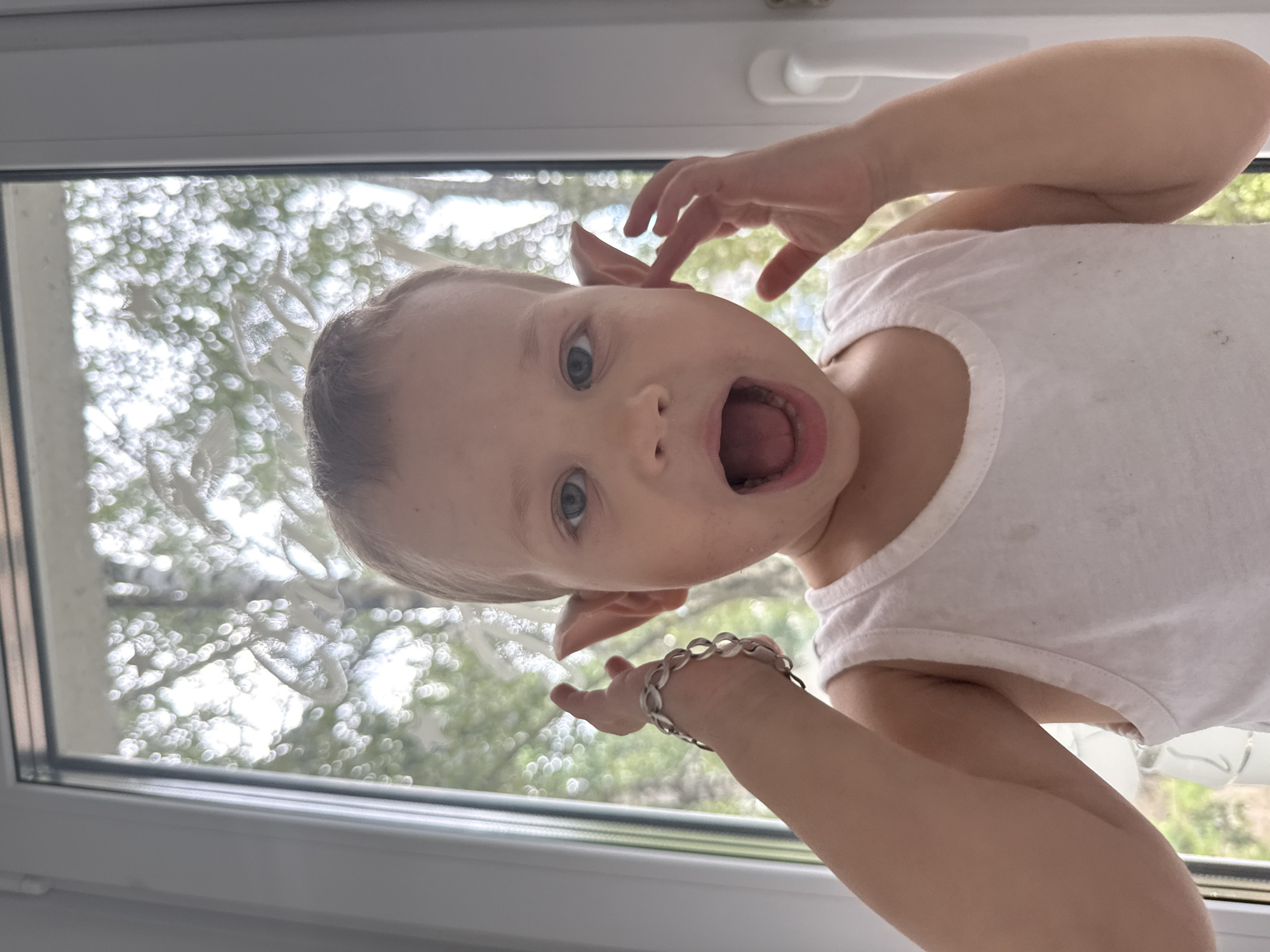 A close-up, chest-up, medium shot captures a fair-skinned, fair-haired toddler with blue eyes against a blurred background of a window with trees outside, possibly in or near Rusanovo, Russia. The child wears a white tank top and has a silver-colored bracelet on their left wrist. Their mouth is wide open in an excited expression, and their hands are positioned near their ears, with the left hand touching their bracelet. The child appears to be mimicking pointed ears or playfully covering their ears. The overall lighting is bright, suggesting daylight.