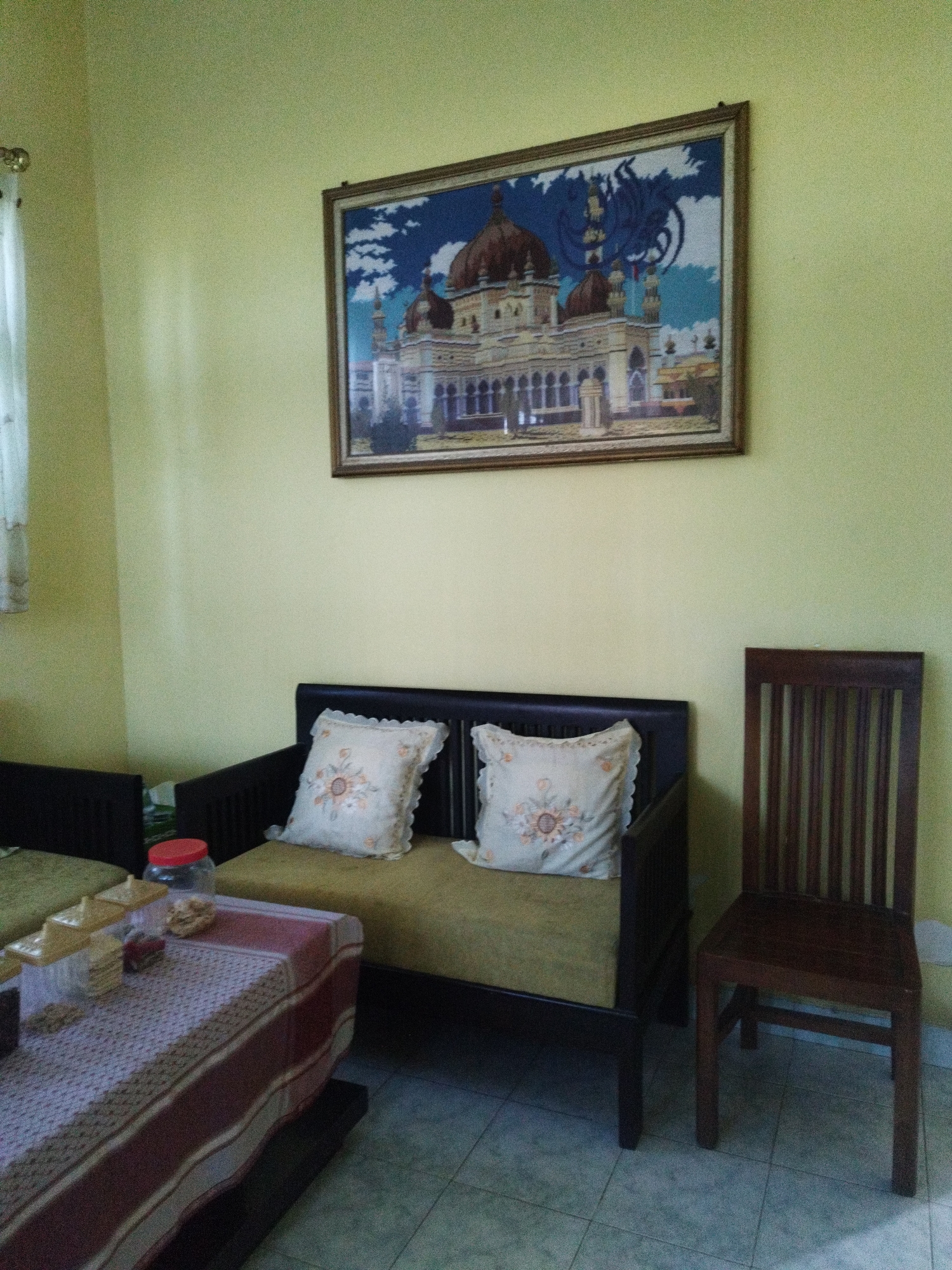 An indoor shot captures a living room area with lime green walls. A large framed tapestry depicting a mosque and a blue dragon-like motif hangs prominently on the wall. In front of the tapestry is a dark-framed sofa with a light green cushion and two decorative pillows. To the right of the sofa, a dark wooden chair with vertical slats stands on a tiled floor. A table covered with a red and white patterned cloth is situated to the left, adorned with several plastic containers and a red-lidded jar. This scene is likely from a residence in Sumenep, Madura, East Java, Indonesia, known for its Islamic cultural heritage and architectural influences, including mosques.
