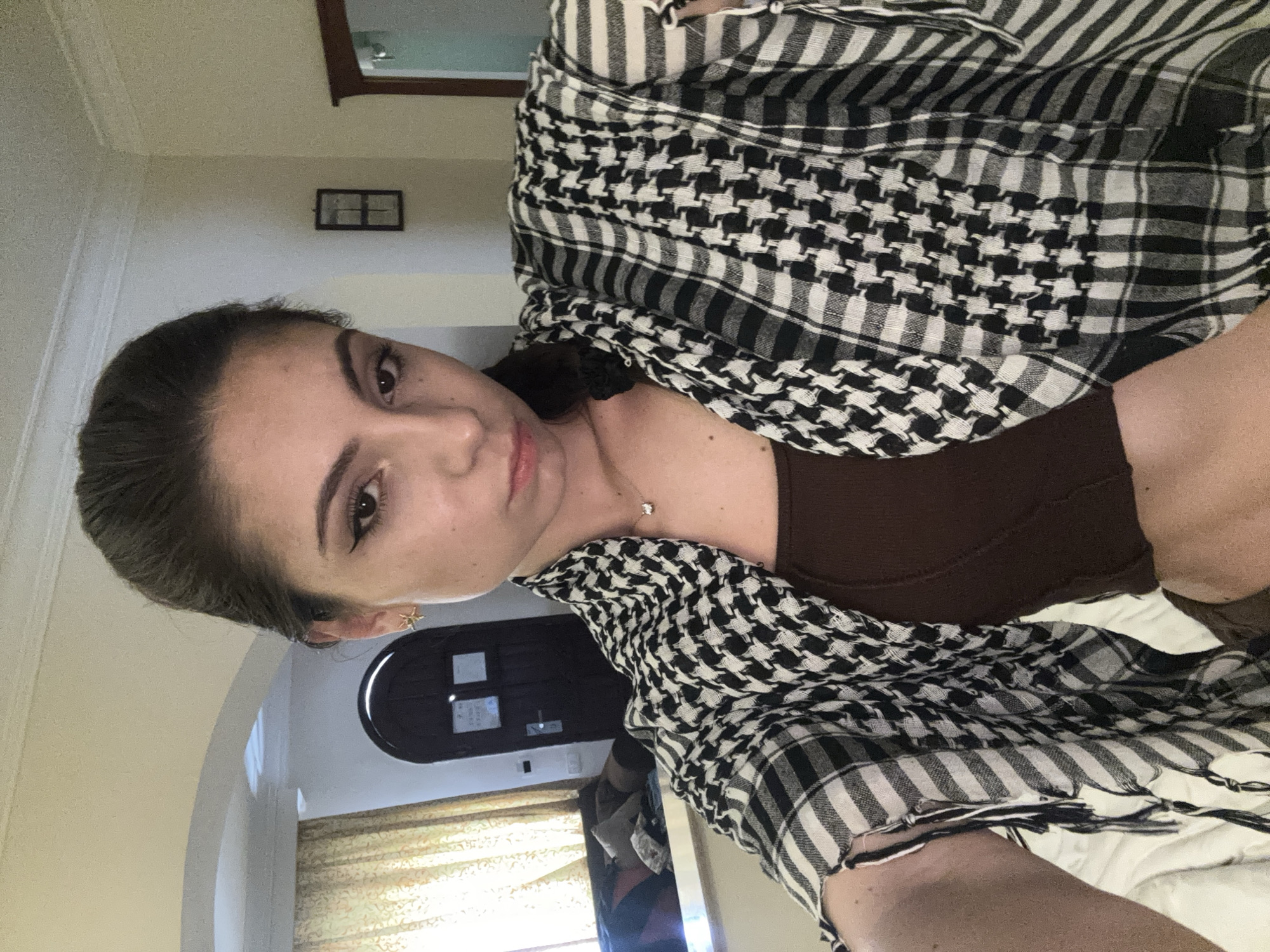 The image is a medium shot of a woman in an interior setting, potentially in the city of Unknown, Unknown. The woman is wearing a brown top and a black and white patterned scarf. She has brown hair and fair skin. She is looking directly at the camera.
Behind the woman, a doorway and a window with curtains are visible. The interior appears to be a room in a home or hotel. A framed picture is on the wall.