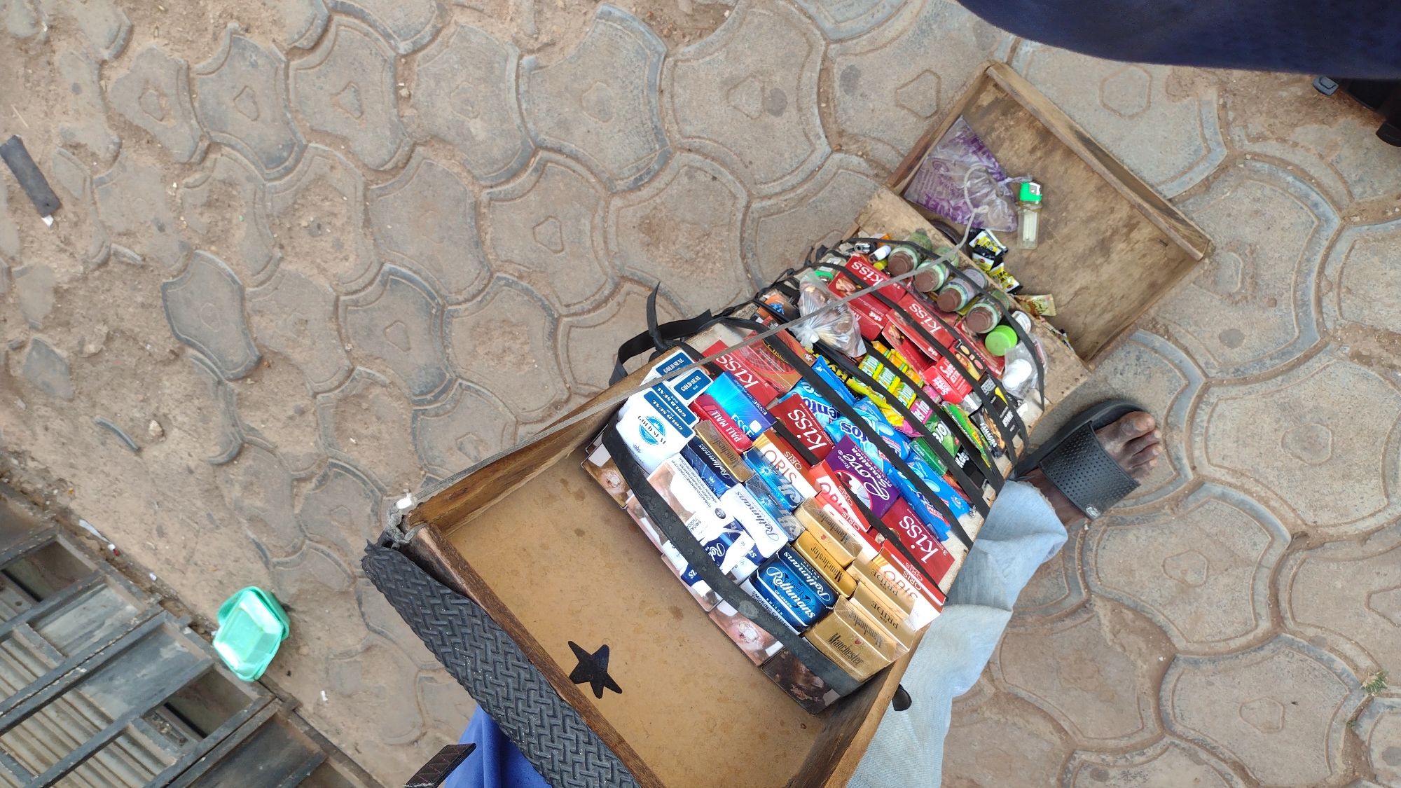 This image depicts a street vendor in Jos, Nigeria, displaying cigarettes and other small items in a wooden case. The vendor, wearing a sandal, is standing on a paved sidewalk. The wooden case is divided into compartments, with the cigarettes arranged by brand. Other items, like small plastic bottles, are also visible. The pavement is made of patterned concrete blocks.