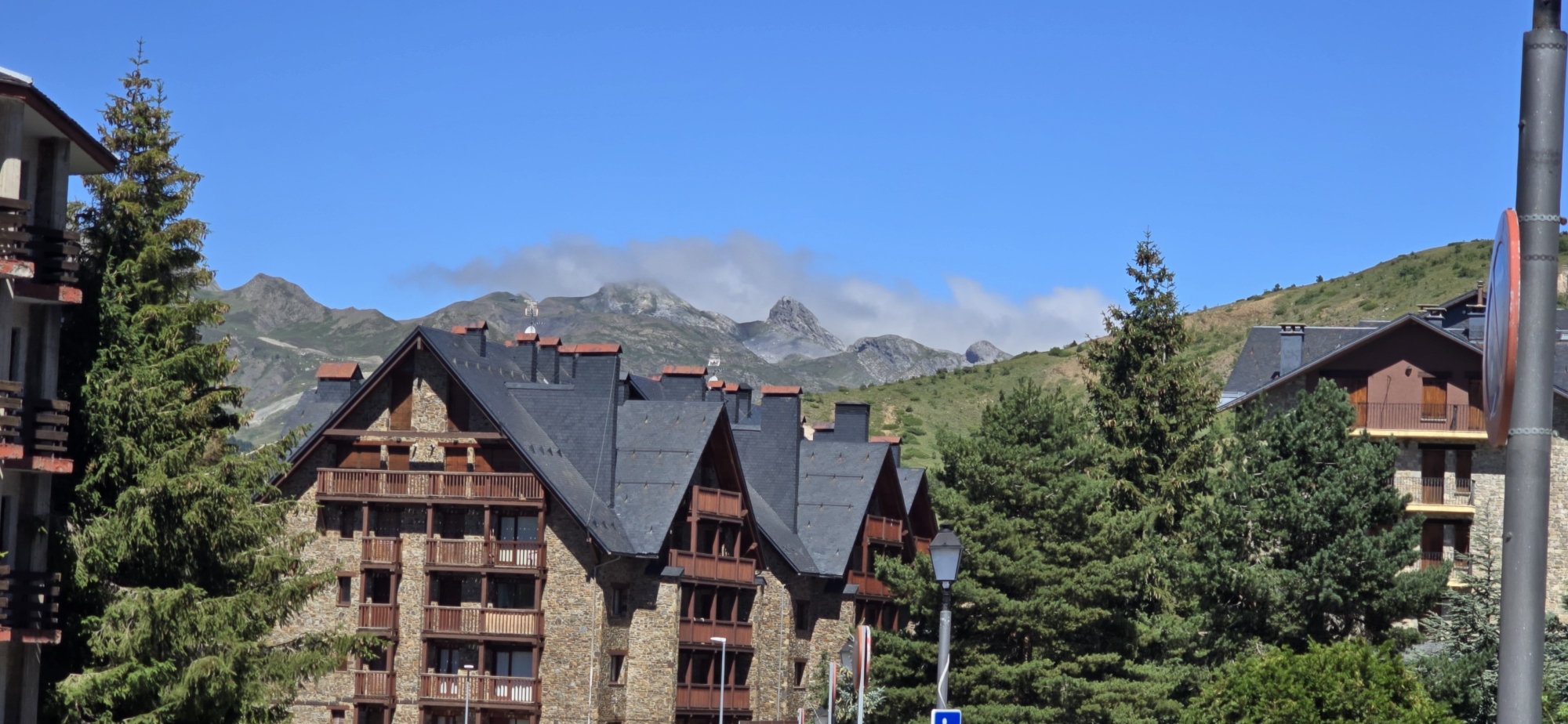 The image depicts a mountain resort setting in Sallent de Gállego, Spain, on a clear, sunny day with a bright blue sky. In the foreground, several multi-story buildings with stone facades and dark, pitched roofs dominate the scene. These buildings feature numerous balconies with wooden railings. Tall evergreen trees, likely pine or fir, are interspersed between the buildings, adding to the alpine atmosphere.
In the background, a range of majestic mountains rises, with some of their peaks partially obscured by wispy white clouds. The lower slopes of the mountains are covered in green vegetation, indicating a summer or early autumn season. The architecture of the buildings suggests a style common in ski resorts, designed to blend with the natural mountainous environment. The overall impression is one of a peaceful, scenic mountain town. There are no people visible in the image, and no actions or interactions are depicted.