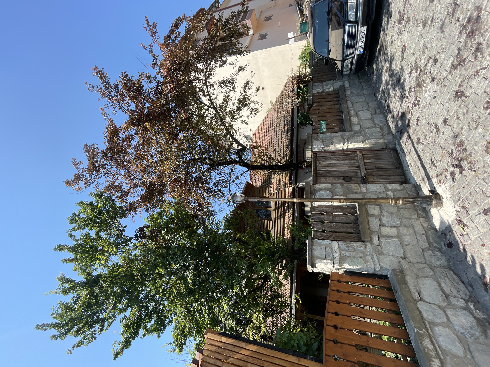 In Mtskheta, Georgia, on a sunny day, a black Mercedes-Benz sedan with license plate LH 360-LH is parked on a cobblestone street in front of a stone and wooden fence. Behind the fence is a traditional-looking house with a red tile roof, partly obscured by two large trees, one with green leaves and the other with reddish-brown leaves. Adjacent to the house is a taller, cream-colored building. A street lamp stands on the sidewalk. Fallen leaves are scattered across the cobblestones.