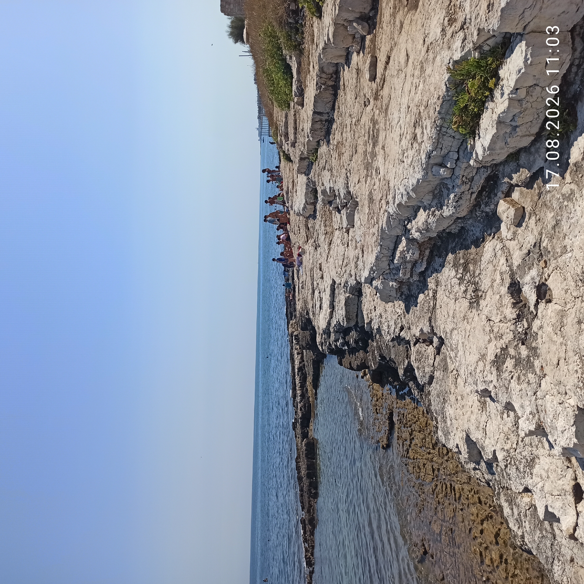 This image depicts a rocky coastline with a body of water and a clear blue sky. In the mid-ground, a group of people are gathered on the rocks, some sitting and others standing. The sea appears calm with small waves lapping at the shore. The coastline itself is characterized by large, light-colored rock formations with some vegetation sparsely growing in the crevices. The overall scene suggests a sunny day at a coastal location. The date "17.08.2026 11:03" is superimposed on the lower right portion of the image, indicating when the photograph was taken. This location is identified as the "Corso Dante" area in Molfetta, Italy.