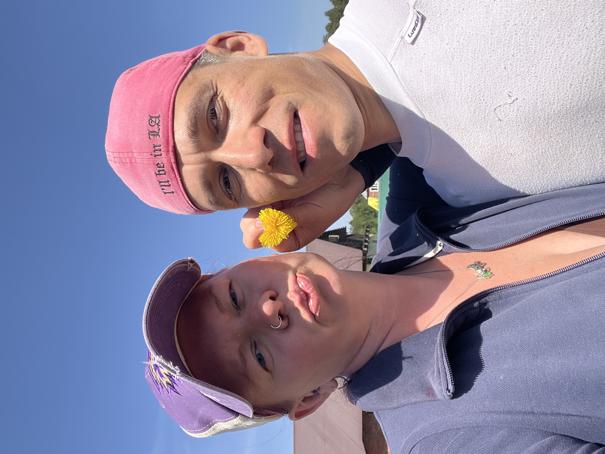 Two individuals are posing outdoors in the daytime with clear blue sky visible. The first person, wearing a pink baseball cap with the text "I'll be in LA" on it, is holding a yellow dandelion flower. They are also wearing a white shirt. The second person is wearing a purple baseball cap and a blue jacket over a low-cut shirt, a necklace with clover charms, and has a nose ring. The location of the image is assumed to be Chaikovsky, Russia.