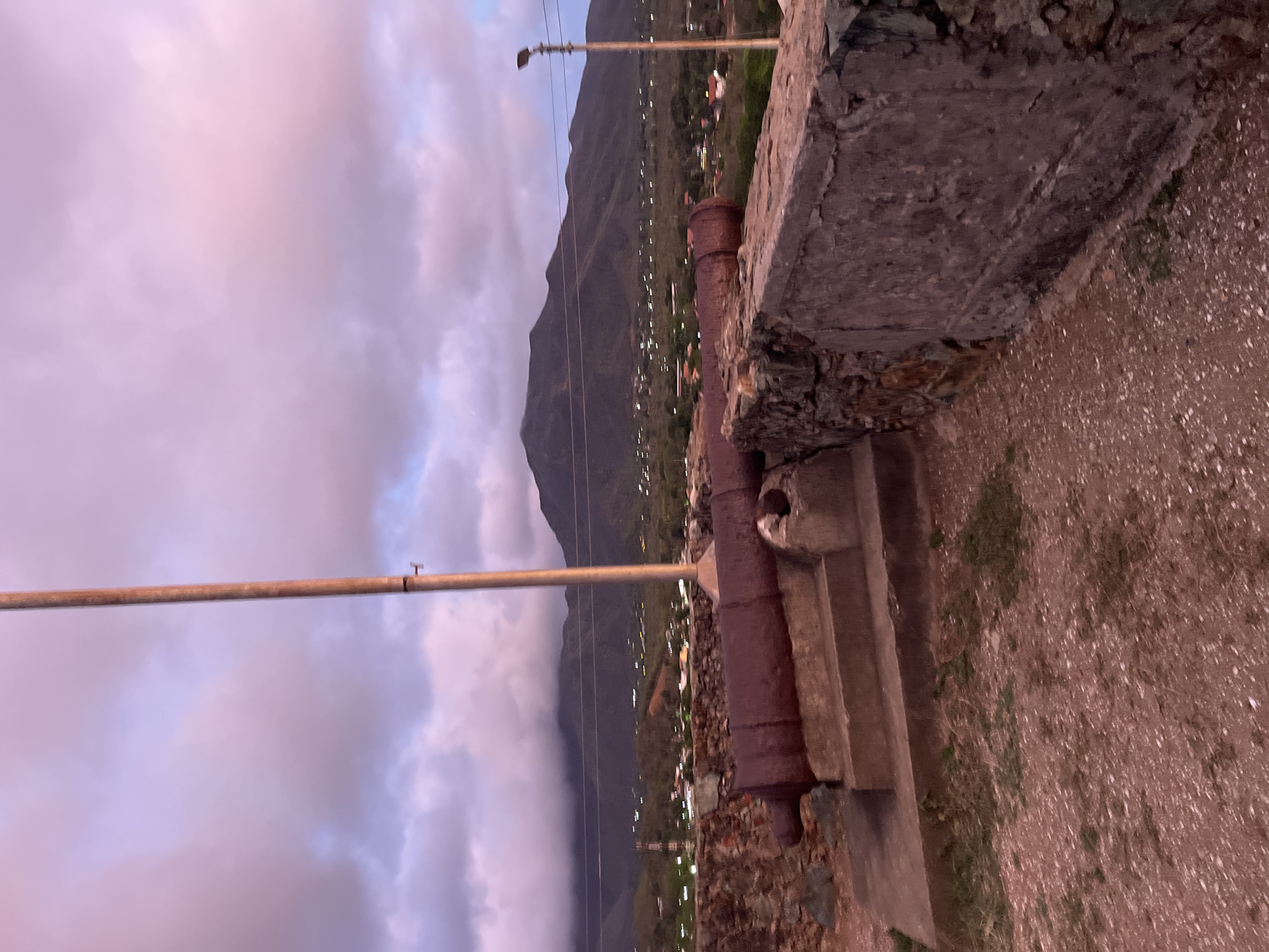 The image depicts a historical scene with two old, rusted cannons positioned on a stone wall overlooking a town and mountains. The sky is filled with dramatic clouds, tinged with the soft hues of either sunrise or sunset. A tall flagpole stands prominently to the right of the frame, its pole casting a long shadow across the scene. The cannons, made of dark, weathered metal, rest on concrete supports, suggesting they are part of a historical defense or monument. The town below is visible with scattered lights, indicating it is either dusk or dawn. The surrounding landscape is mountainous, with a range of hills forming a backdrop to the town. The foreground consists of a rough, gravelly surface with some sparse vegetation. Based on the presence of these cannons and the historical context associated with them, this location is likely Fort Barrancas, a historical site in Santa Ana, Venezuela, known for its defensive artillery. No people are visible in the image, and the overall mood is one of quiet historical reflection.