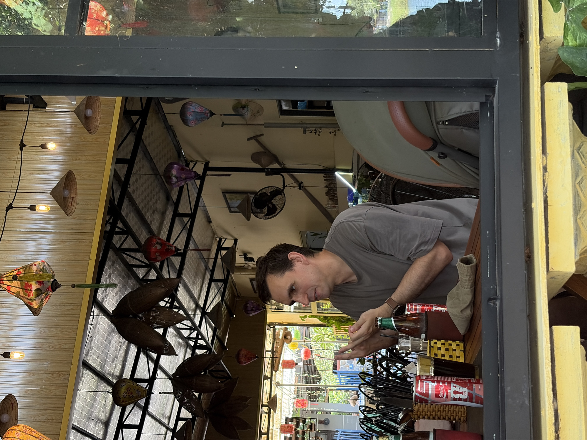 The image captures a man seated at a wooden table inside a cafe or restaurant, viewed from an exterior perspective through a window or open frame. The man, with dark hair and wearing a grey t-shirt and a watch on his left wrist, is looking downwards, seemingly focused on an object in his hands. He is holding a green glass bottle with his left hand.
On the table in front of him, there is a brown and yellow checkered placemat, a light brown fabric object (possibly a boot or a container), and a silver and red can of Coca-Cola with "FIFA" branding clearly visible. Another green glass bottle, likely containing beer, is also on the table.
The interior of the establishment features a wooden slat ceiling adorned with numerous traditional Vietnamese lanterns. These include conical hat-shaped light fixtures, intricate woven bamboo lanterns, and colorful fabric lanterns of various designs and sizes. Several ceiling fans are also visible. Through the window or opening, a bright outdoor scene can be observed, featuring green foliage, architectural elements, and more hanging lanterns in vibrant red, yellow, and blue colors. The natural light suggests it is daytime. The man's expression is neutral and concentrated. The setting is consistent with a cafe or restaurant in Hội An, Vietnam, given the distinct style of lanterns.