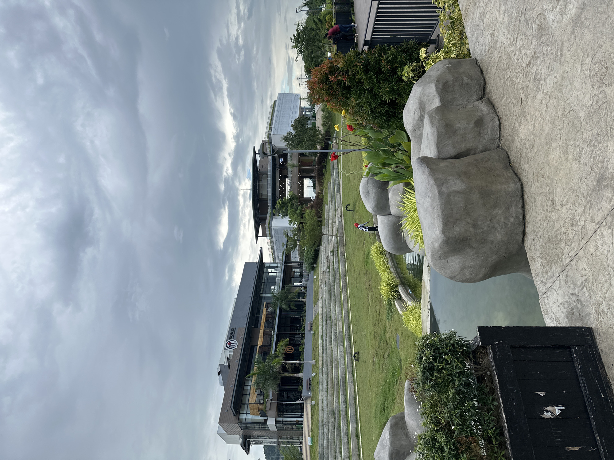The image depicts the Azuela Cove in Davao City, Philippines, on an overcast day. In the foreground, there is a paved walkway with decorative concrete texture, adjacent to a shallow pond surrounded by large, gray, artificial rocks and lush green plants. A black planter box with a white bush is visible to the left. Beyond the pond, a tiered grassy area with concrete steps leads up to a modern, multi-story building with a dark facade and large glass windows. A sign with a logo featuring a circular emblem with the letters "FI" is visible on the building. To the right of this building, another modern structure with white decorative panels and glass windows is visible. Several palm trees and other greenery are scattered throughout the landscape. In the middle ground, a person in a red and white outfit appears to be tending to the plants or grounds. In the background, a few other individuals are visible near a railing, suggesting public access to the area. The sky is filled with heavy, gray clouds, contributing to a subdued lighting condition.