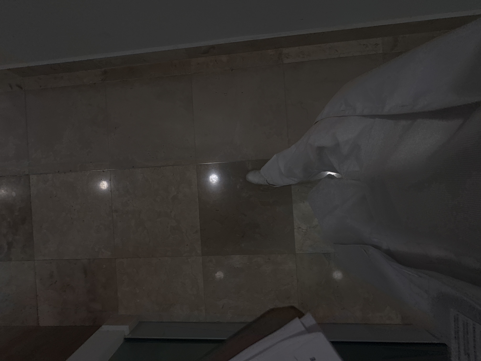 An overhead shot of a tiled floor, partially covered by a white garment or sheet. A white-clad person is partially visible towards the right of the frame, near the garment. Paper or documents are in the lower portion of the image. This photo was taken in Puebla, Mexico.