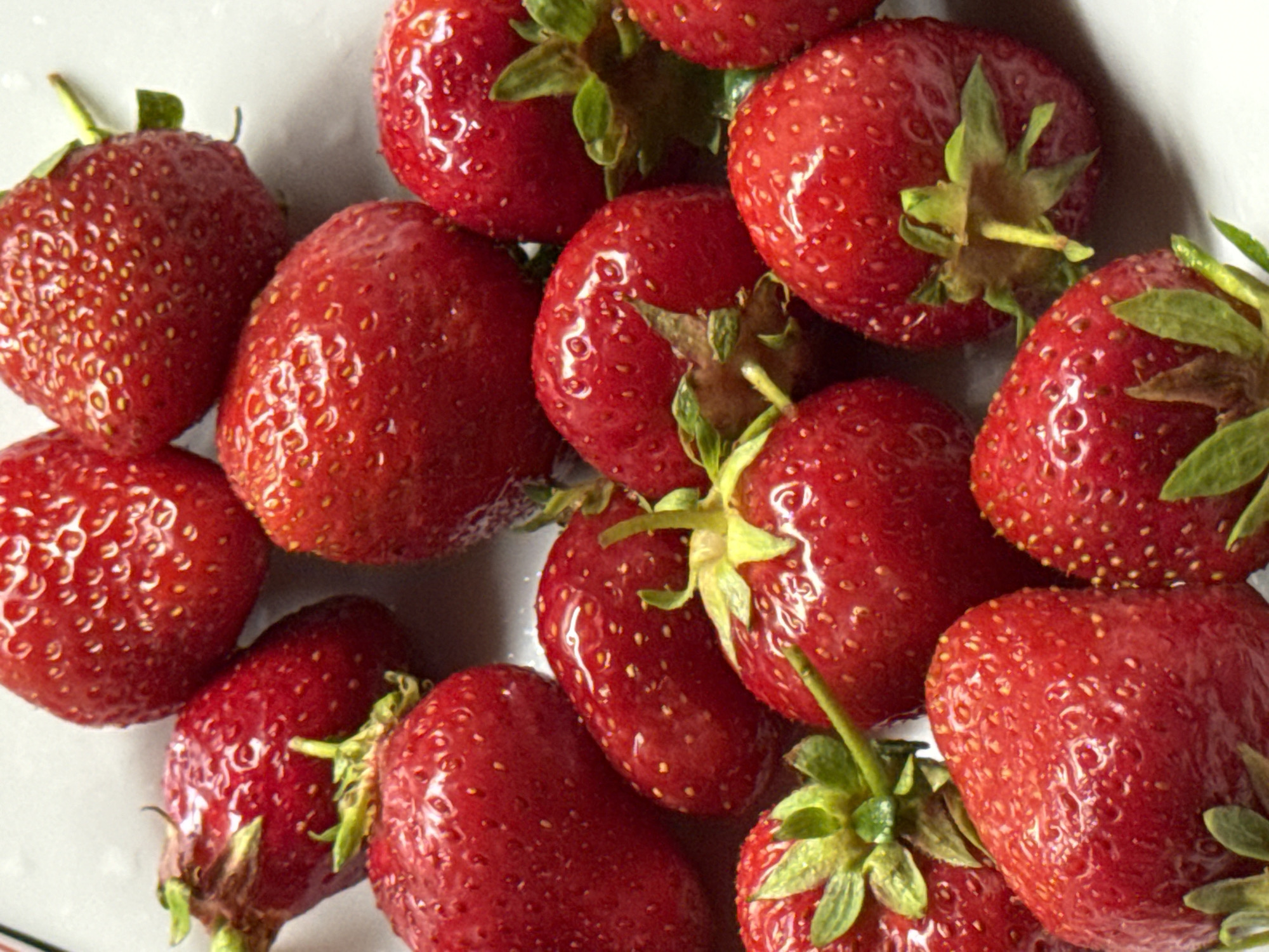 A close-up, overhead shot captures a pile of ripe strawberries arranged on a white surface. The strawberries are a vibrant red, with visible seeds dotting their surfaces and green stems peeking out from some. The lighting highlights the glossy texture of the fruit, creating subtle reflections. The background is out of focus, suggesting a shallow depth of field. There are no people, actions, or interactions depicted in the image. The image does not provide any information about the location of Budva, Montenegro.