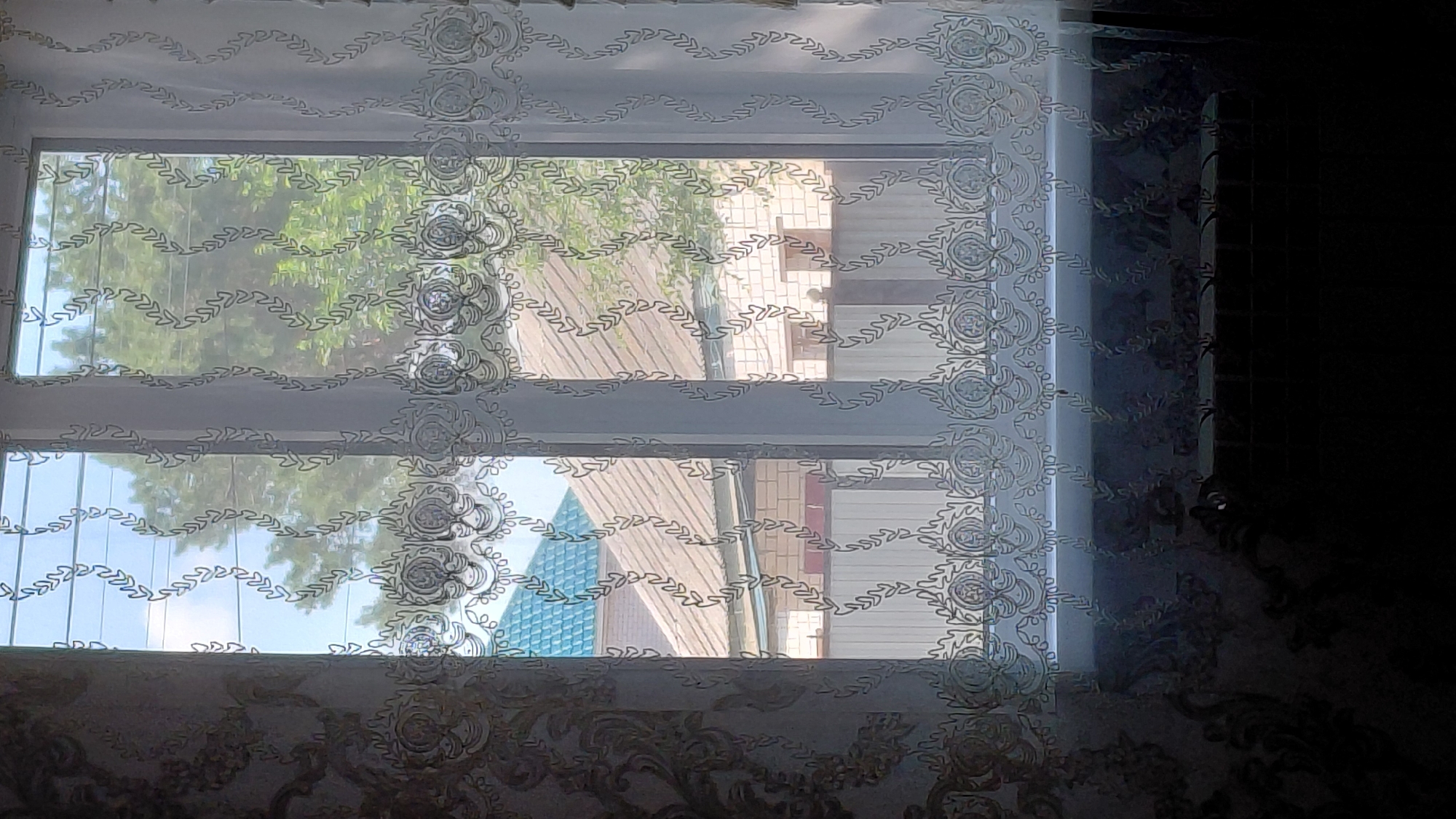 The image depicts a window with white, semi-sheer curtains featuring an embroidered floral vine pattern along the bottom and a more elaborate, repeating circular motif above. The curtains are drawn, revealing a view of residential buildings and trees outside. One building has a teal-colored roof, while others have corrugated metal roofs and brick facades. A white fence is also visible. The interior of the room is dimly lit, with a dark, patterned wallpaper on the left and a dark area across the bottom of the frame, possibly a radiator or furniture. The overall impression is that of a residential interior looking out onto a suburban or semi-urban landscape. The mention of Aktobe, Kazakhstan suggests this is a domestic scene within that specific geographic location.