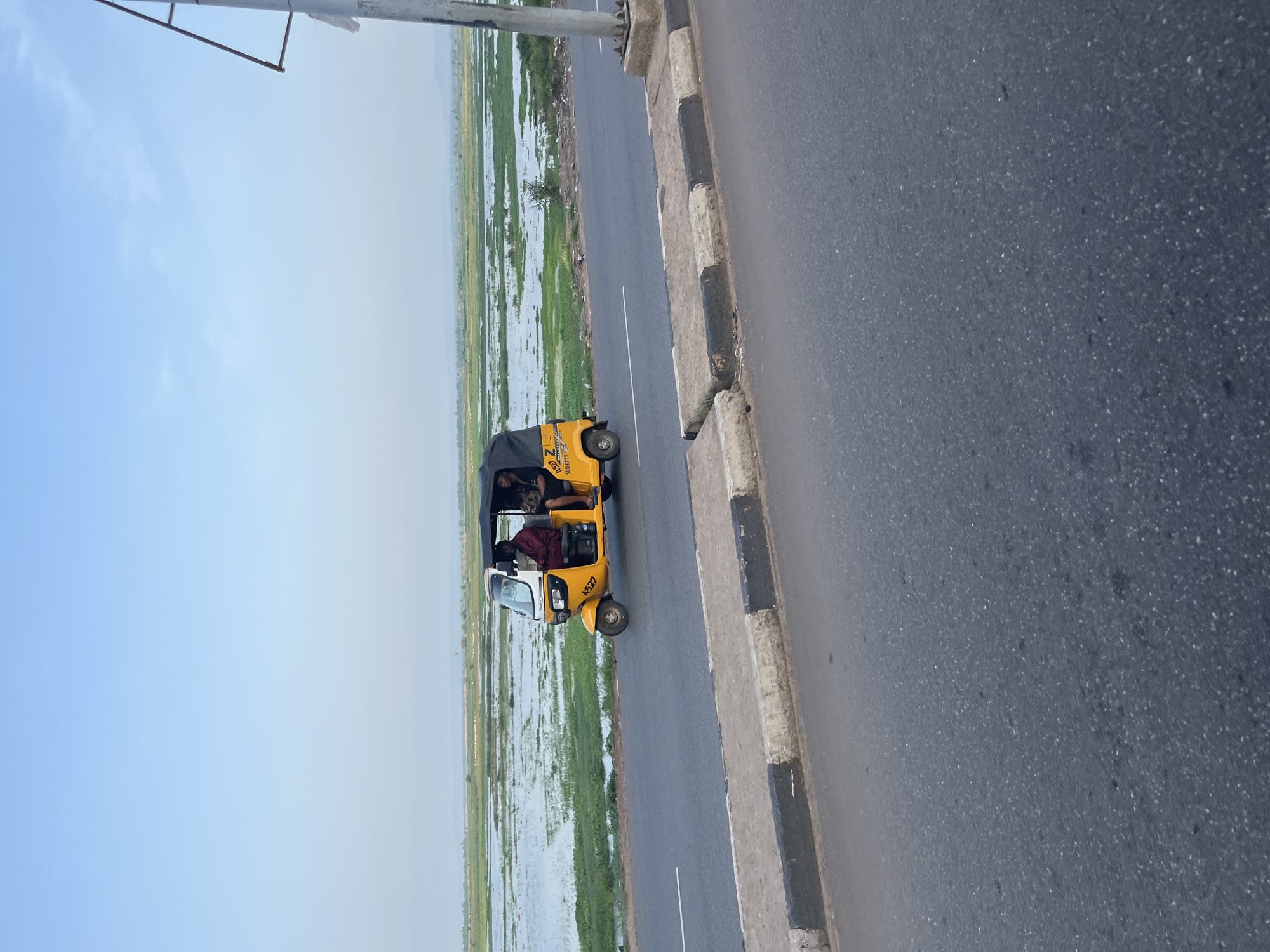 A yellow auto-rickshaw travels on an asphalt road. The auto-rickshaw has a black canopy and is carrying two passengers. The road is bordered by a concrete curb with black and white markings. In the background, there is a body of water with green vegetation surrounding it, and a hazy horizon with distant hills under a pale blue sky. The scene appears to be in an outdoor environment, possibly a rural or semi-rural area.
