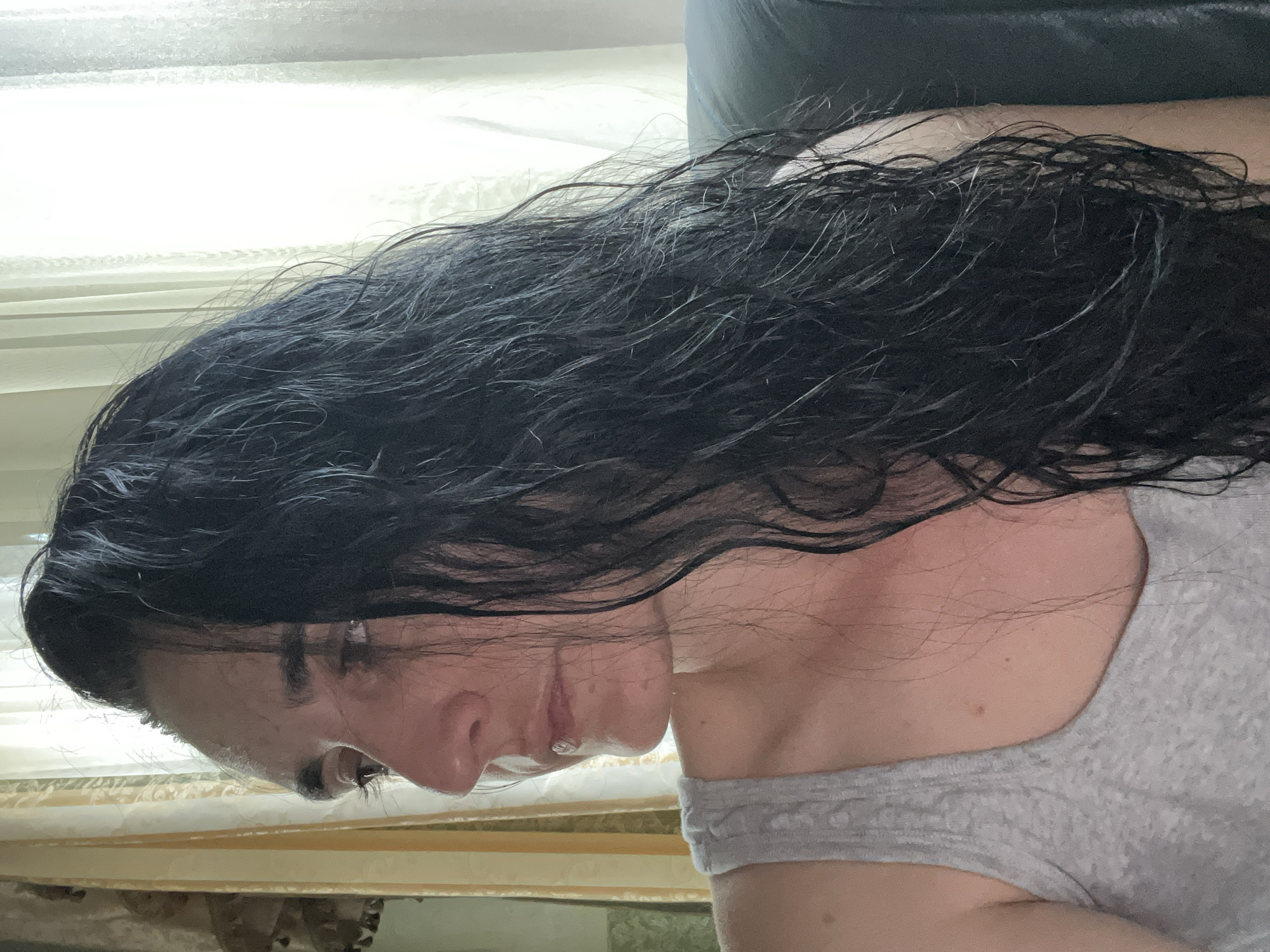 A female subject is depicted in a left-side profile, positioned indoors within the city of Tarnivtsi, Ukraine. She possesses long, dark, wavy hair that appears wet or damp, with discernible individual strands. The subject is dressed in a light gray, ribbed sleeveless top. Her gaze is directed towards the left, off-frame. The immediate background on the right consists of brightly illuminated, sheer white curtains covering a window, indicating a source of natural light. To the left, striped yellow and white drapes are visible, alongside a partial view of a decorative tassel. The subject's skin exhibits some reddening on the visible neck and upper chest area. She appears to be seated, with a dark-colored furniture back visible in the lower right portion of the frame.