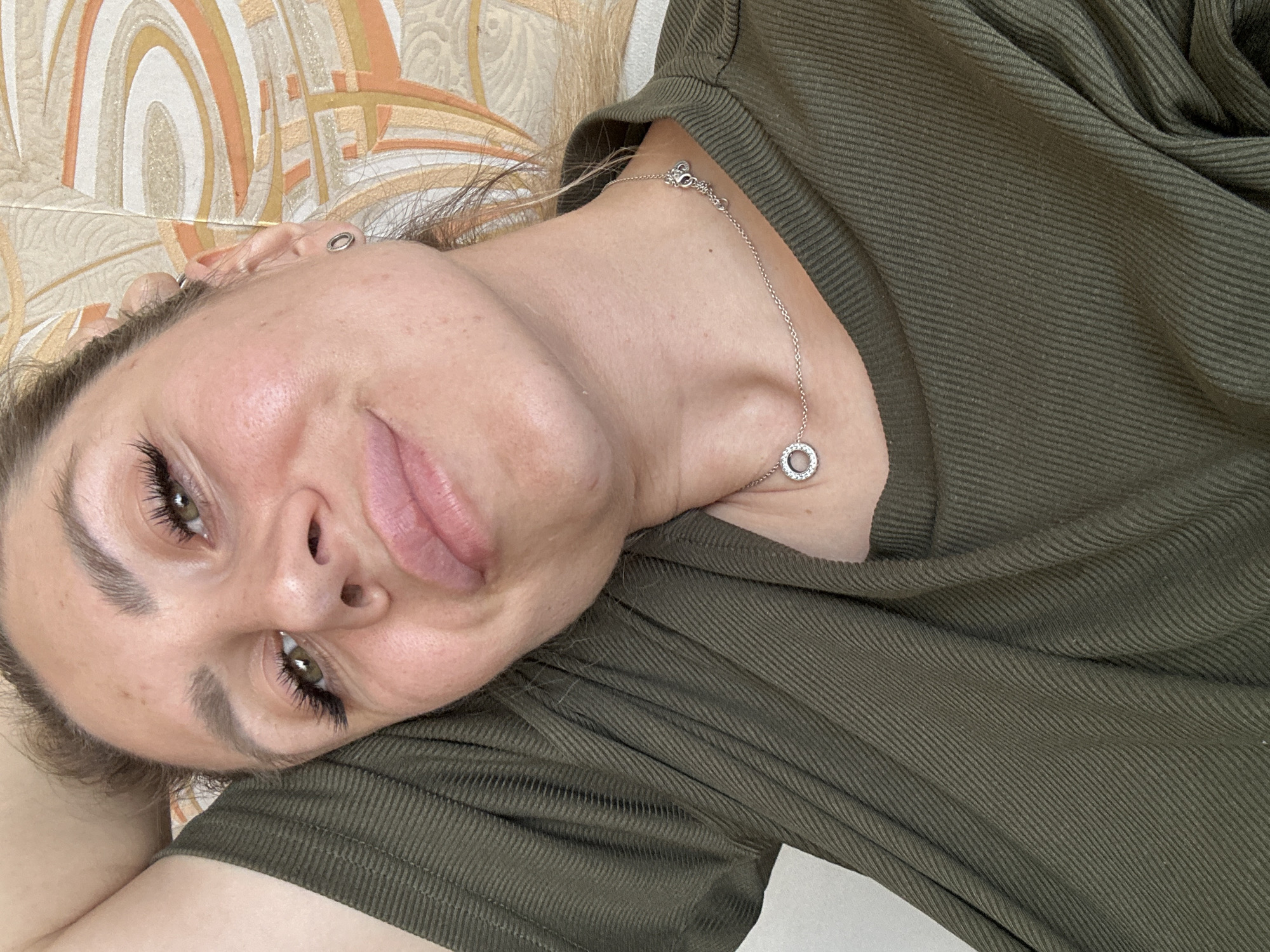 A close-up, overhead shot shows a woman with light brown hair and green eyes wearing a green, ribbed t-shirt and a silver necklace. Her left arm is bent behind her head, supporting it as she reclines. Her head is tilted slightly to the right, and she is looking directly at the camera with a neutral expression. Her lips are slightly parted. The background consists of a wall with a beige, abstract pattern. The lighting is natural and appears to be coming from the right. There are no visible landmarks or elements that would indicate the specific location within Novoaltaysk, Russia.