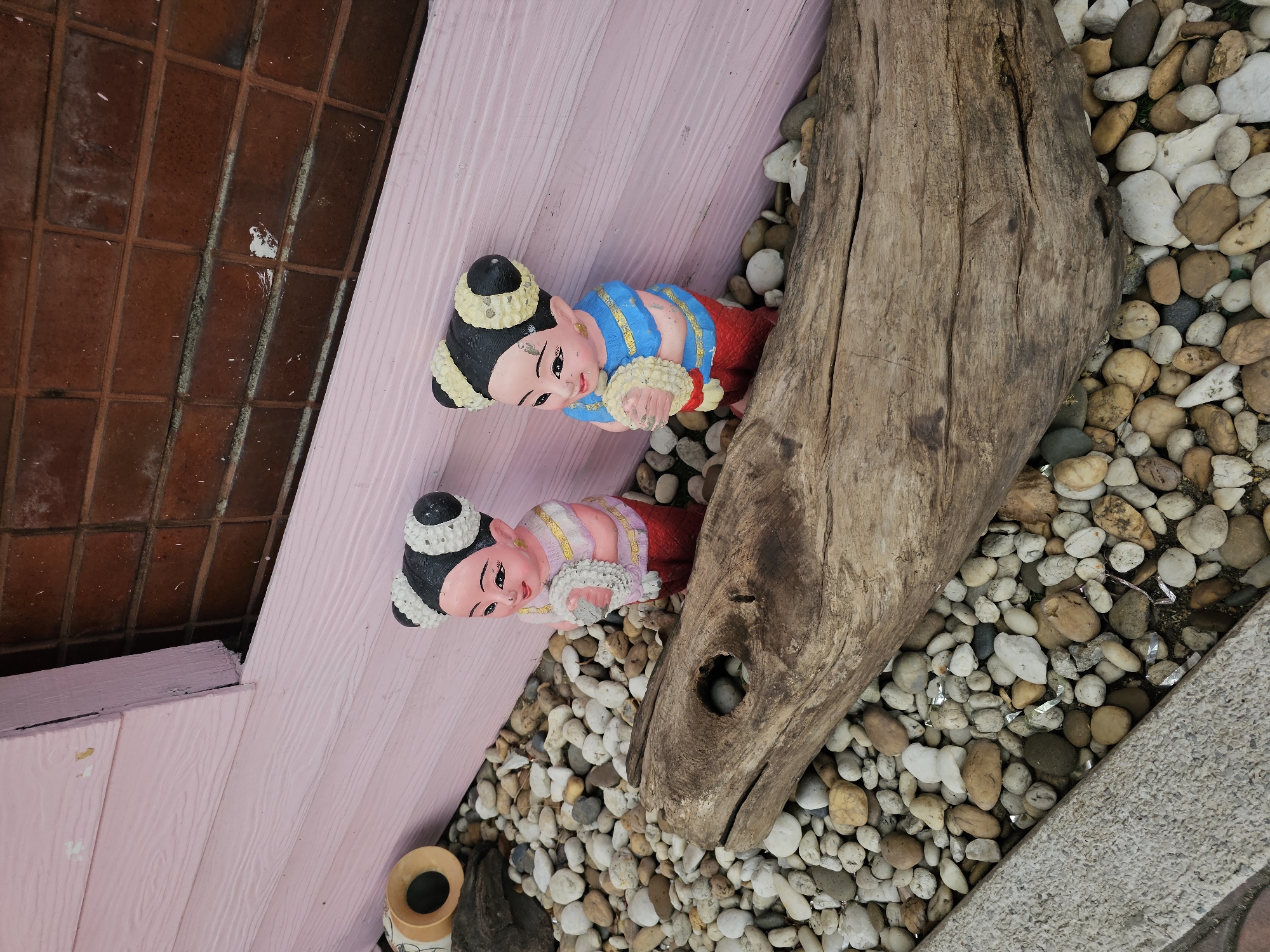 The image depicts a close-up, outdoor view of two decorative ceramic figurines of young girls, positioned on a bed of river stones. The figurines, which appear to be in traditional attire, are situated in front of a pink, wooden-paneled wall and a brick wall. A large piece of weathered driftwood lies horizontally in the foreground, partially obscuring the figurines and stones. To the left of the figurines, a small terracotta pot or planter is visible. The overall scene suggests an outdoor decorative arrangement, possibly in a garden or near the entrance of a building. The setting is consistent with various establishments in Bangkok, Thailand, that utilize such decorative elements to enhance their aesthetic appeal, such as cafes, guesthouses, or small shops.