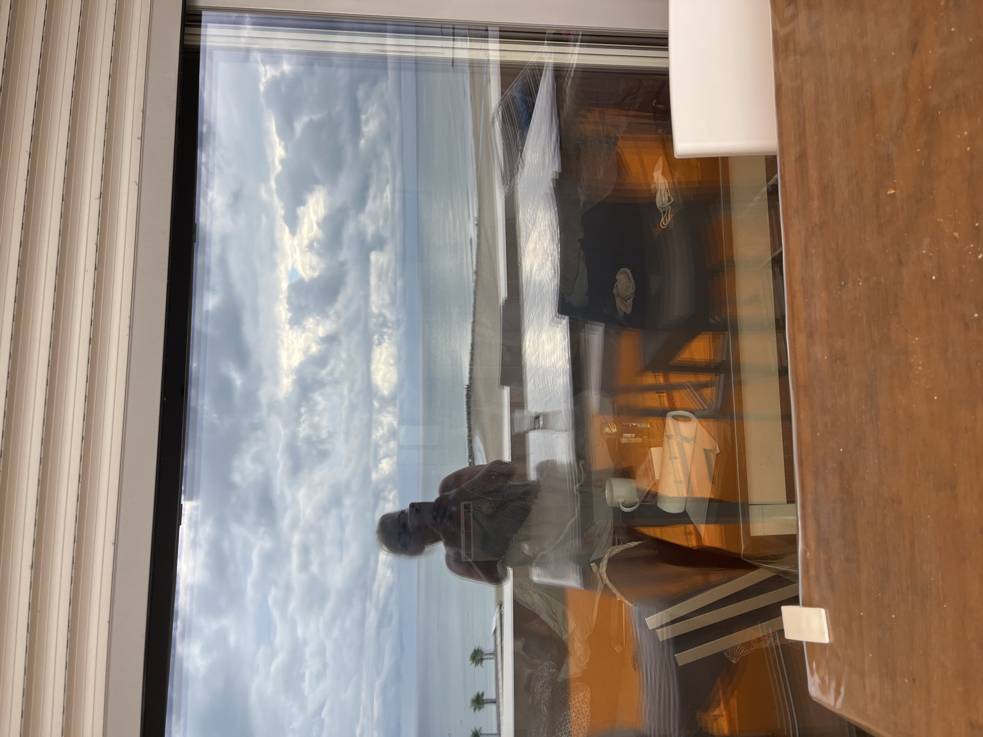 The image captures a reflection in a large window overlooking the beach and sea in Palavas-les-Flots, France. In the reflection, a person is visible taking a selfie with a smartphone. The person appears to be standing on a balcony or terrace. In the foreground, there are reflections of indoor furniture, including a wooden table and chairs with orange upholstery. The sky is cloudy, and the sea is calm. The overall impression is that of a relaxed, indoor setting with a view of the coastline.