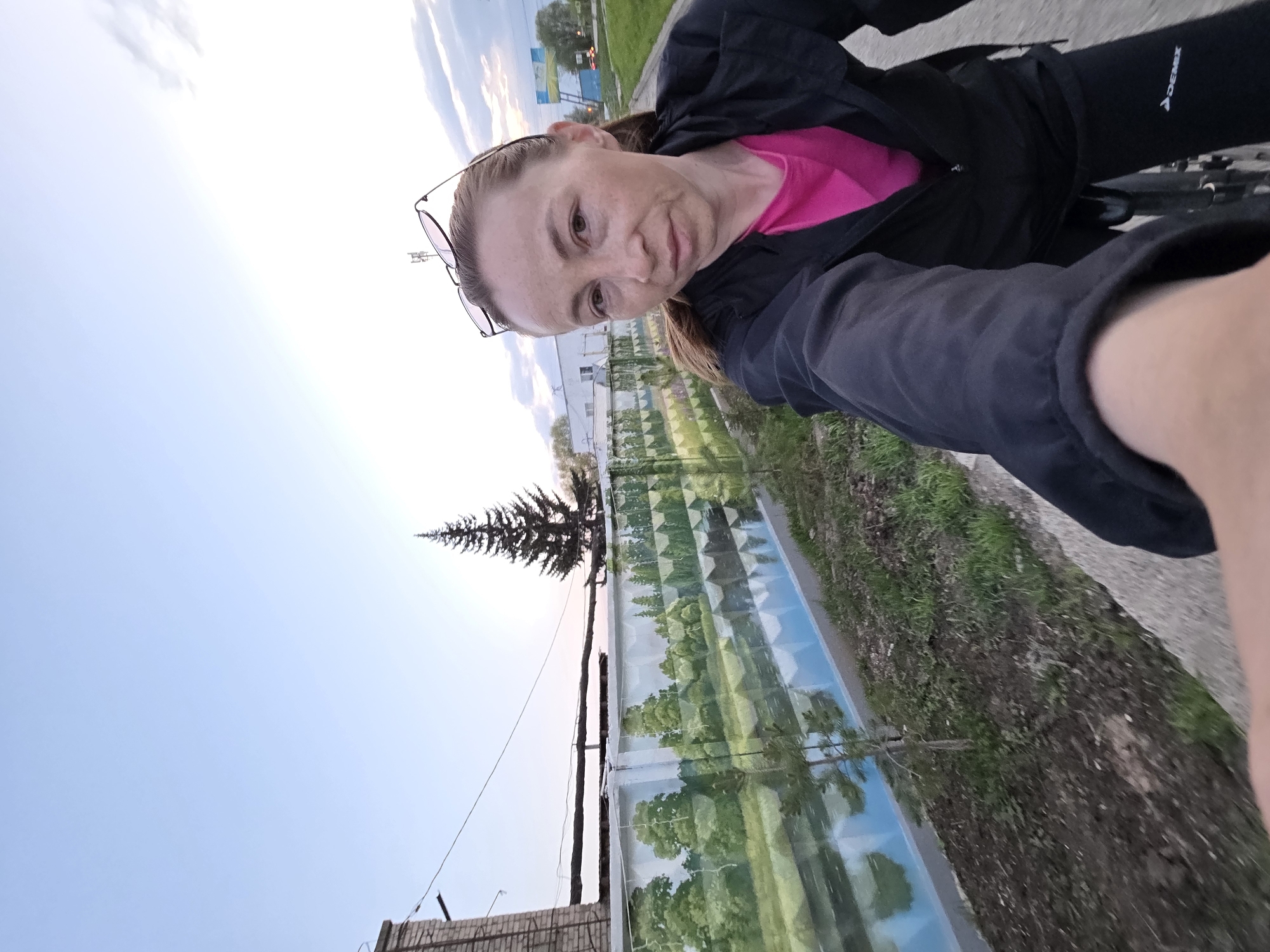 A selfie captures a woman on a bicycle in Chaikovsky, Russia. The woman has glasses atop her head, wears a pink shirt under a black jacket, and is likely wearing black leggings. The background features a long, reflective wall with a scenic mural of trees and sky, juxtaposed against a real evergreen tree. Part of a brick structure is visible in the lower-left corner, contrasting with the expanse of the clear sky. The setting seems to be a paved area, possibly a bike path, with some vegetation visible on the ground.