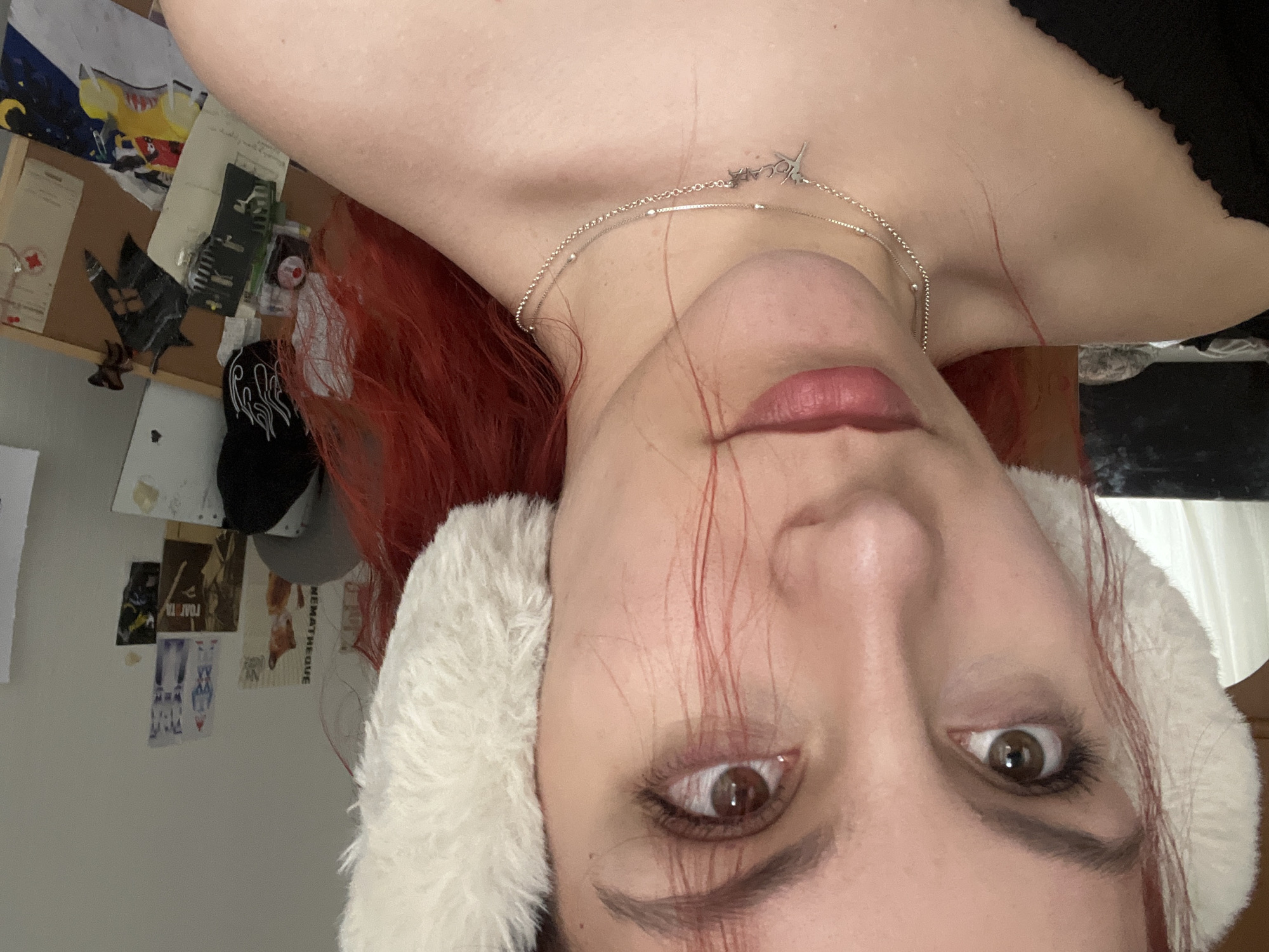 A close-up, inverted selfie depicts a woman with fair skin, brown eyes, and red hair. She is wearing fluffy white earmuffs and a silver necklace. Traces of red hair lie across her face. The background reveals a wall with various pinned items and a window with curtains. The location in Sofia, Bulgaria is undetermined.