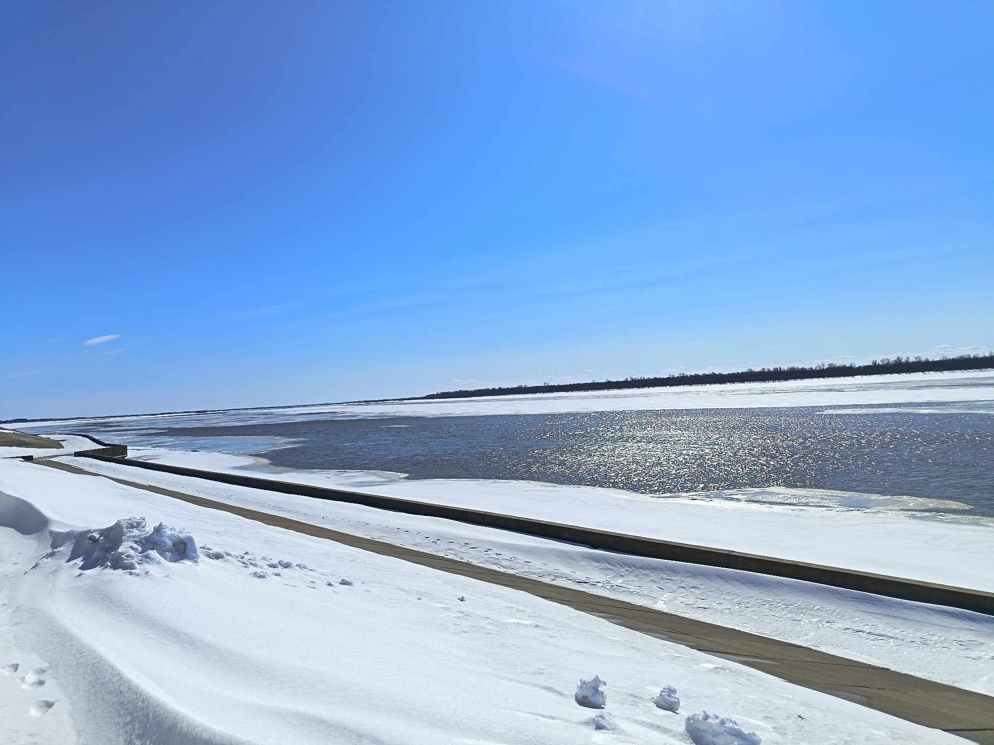 The image depicts a partially frozen riverbank in Nizhnevartovsk, Russia, on a sunny day. The sky is clear blue with minimal clouds. The foreground is covered in snow, with a concrete embankment running parallel to the river. The river has open water with visible sunlight reflecting off the surface, interspersed with areas of ice. In the distance, the opposite bank of the river is visible, lined with trees. The scene suggests a cold, winter environment transitioning towards spring. There are snow drifts on the embankment and patches of bare ground visible. Footprints are visible in the snow, suggesting recent human activity.
