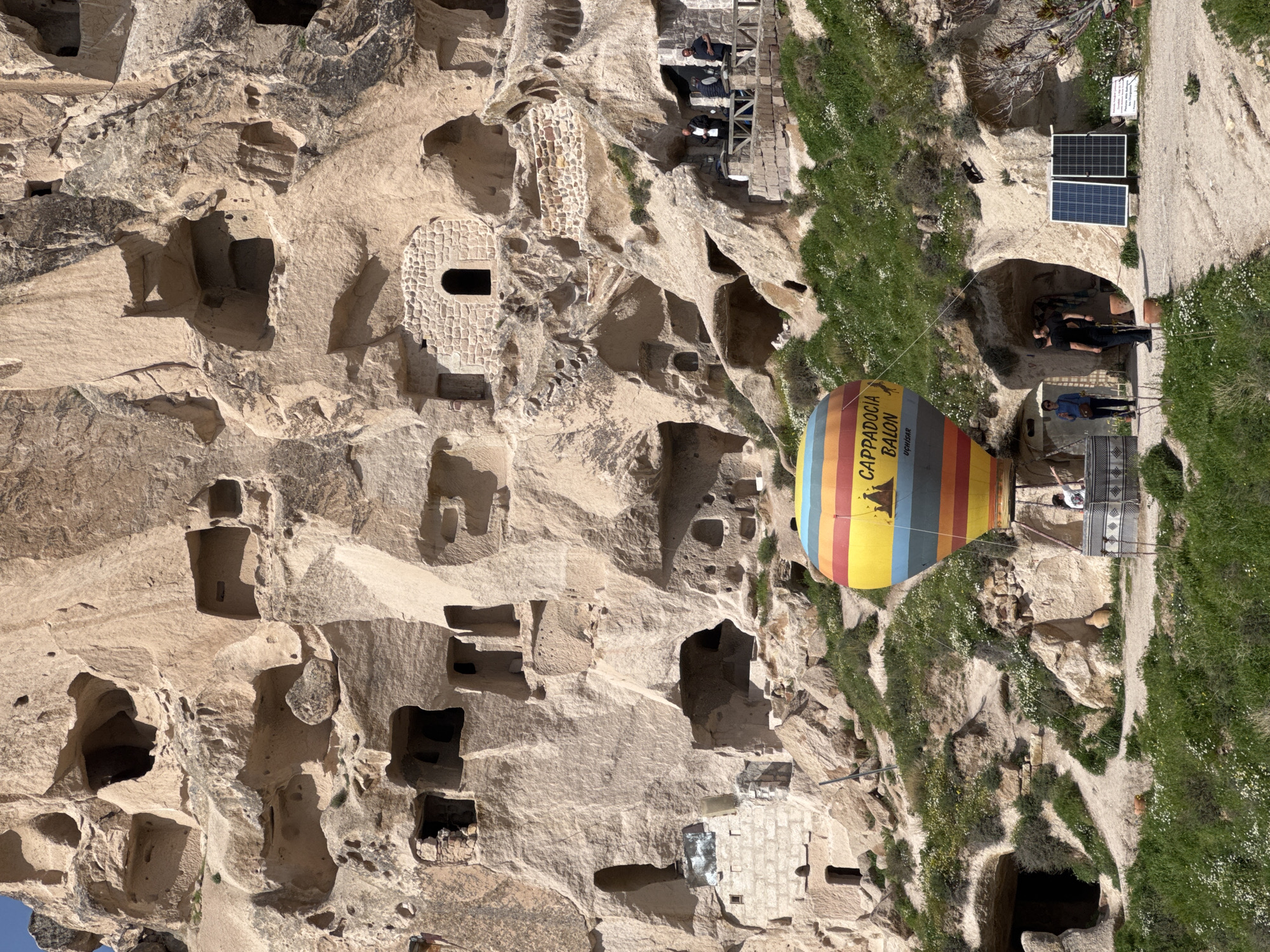 The image depicts a vertical view of a unique landscape in Uçhisar Beldesi, Turkey. The foreground and the left side of the frame are dominated by a light beige, rocky cliff face. This cliff is riddled with numerous dark holes or cave entrances, some appearing as simple openings, while others are more structured with stone facades.
To the right of the cliff face, a vibrant hot air balloon is visible. It features colorful horizontal stripes of yellow, orange, red, blue, and green. The words "CAPPADOCIA BALON" and "UÇHISAR" are printed on it.
Adjacent to the balloon, a stone staircase leads to an upper level. Three individuals stand near the staircase, engaged in conversation. Beyond them, two solar panels are placed on the ground. A dirt path extends upwards, bordered by green grass and scattered white flowers. The environment is a mix of rugged rock formations and verdant vegetation, creating a stark contrast between the natural and man-made elements. The lighting suggests a sunny day, highlighting the textures of the rocks and the colors of the balloon.