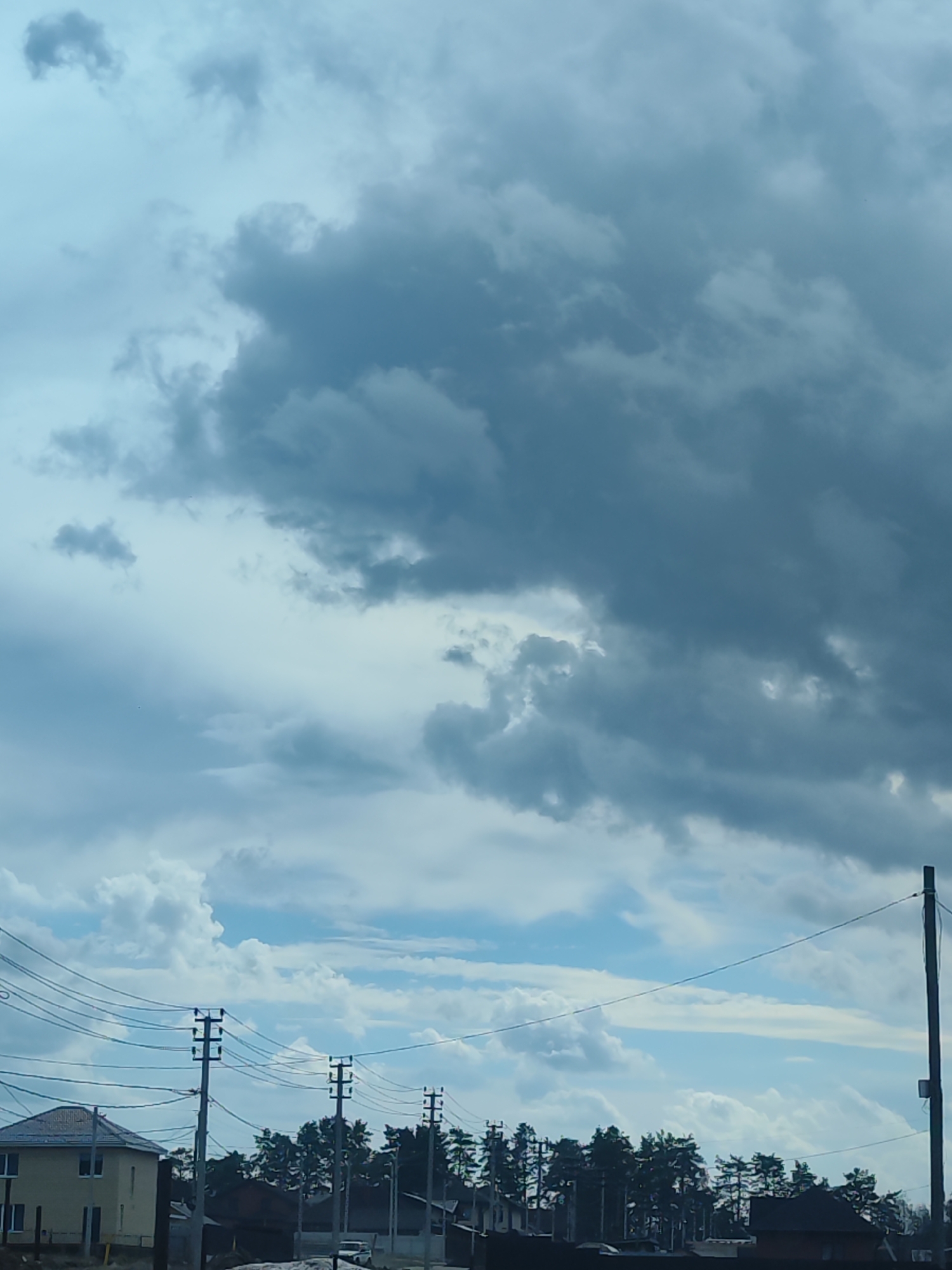 The image displays an outdoor scene dominated by a heavily clouded sky, suggestive of unsettled weather in a residential or semi-rural area of Chaikovsky, Russia. The sky occupies the majority of the frame, filled with dense, dark grey clouds interspersed with brighter patches of blue sky and lighter, white clouds.
In the lower third of the image, the landscape features human infrastructure and natural elements. Several dark utility poles are prominent, supporting a complex network of overhead electrical wires stretching across the scene. To the far left, a light-colored house with a dark-pitched roof is partially visible. A dense band of evergreen trees, likely pine or fir, runs horizontally across the middle ground, separating the structures from the sky and partially obscuring other buildings behind them. A light-colored vehicle, possibly a car or van, can be faintly seen behind the trees on the right side.
The scene is static with no discernible human activity. The time of day is daytime, though the overcast conditions create a diffuse light. There is no visible text in the image. The heavy cloud cover suggests either impending rain, recently passed rain, or simply an overcast day.