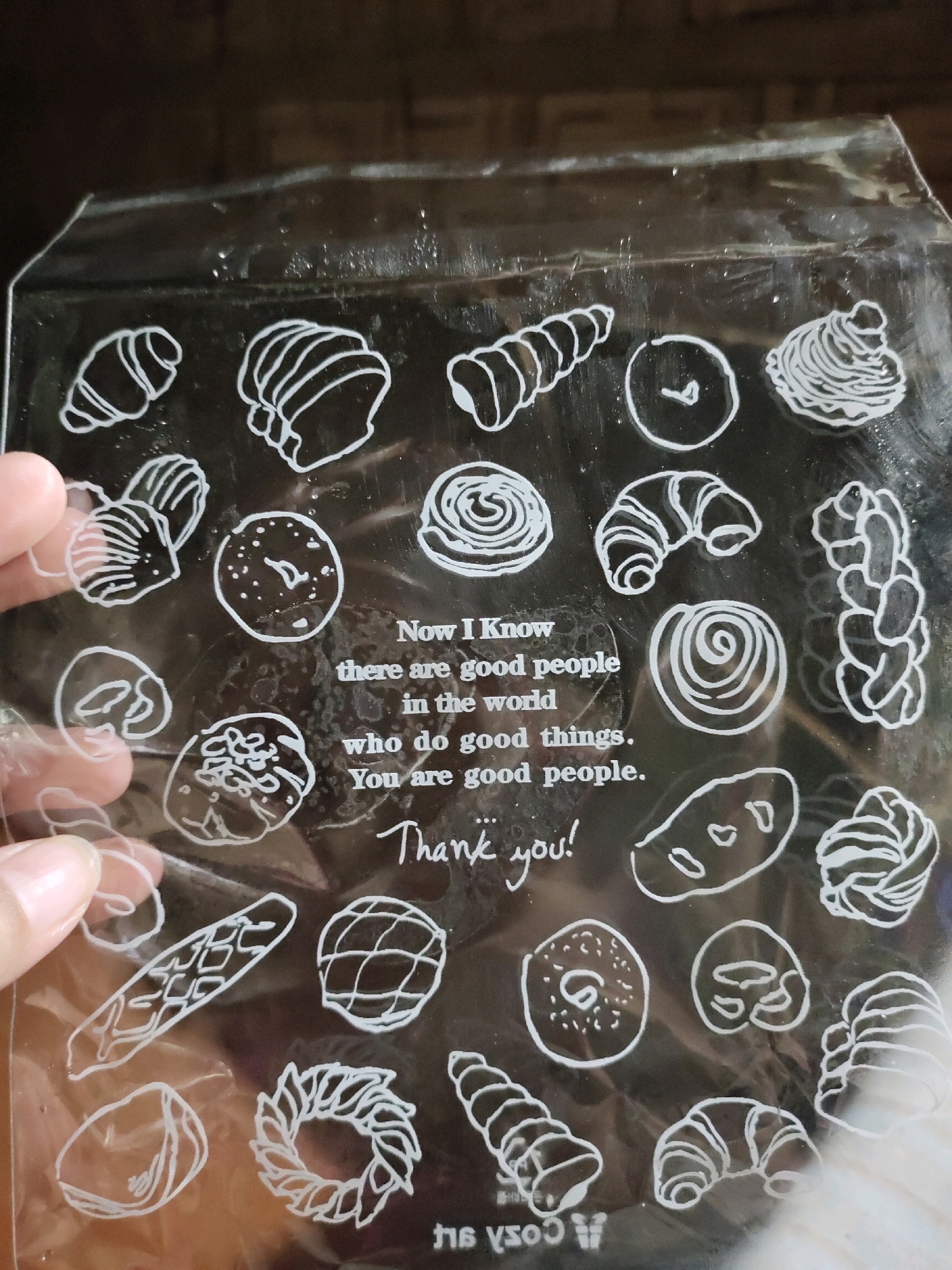 The image shows a clear plastic sheet with white line drawings of various baked goods and text. The baked goods include croissants, buns, bread loaves, a braided bread, and a donut. The text reads: "Now I Know there are good people in the world who do good things. You are good people. Thank you!". A person's hand is visible holding the sheet from the left side. The background appears to be a dark, textured surface, possibly wood. The image does not provide any information about the location of Matalam, Philippines.