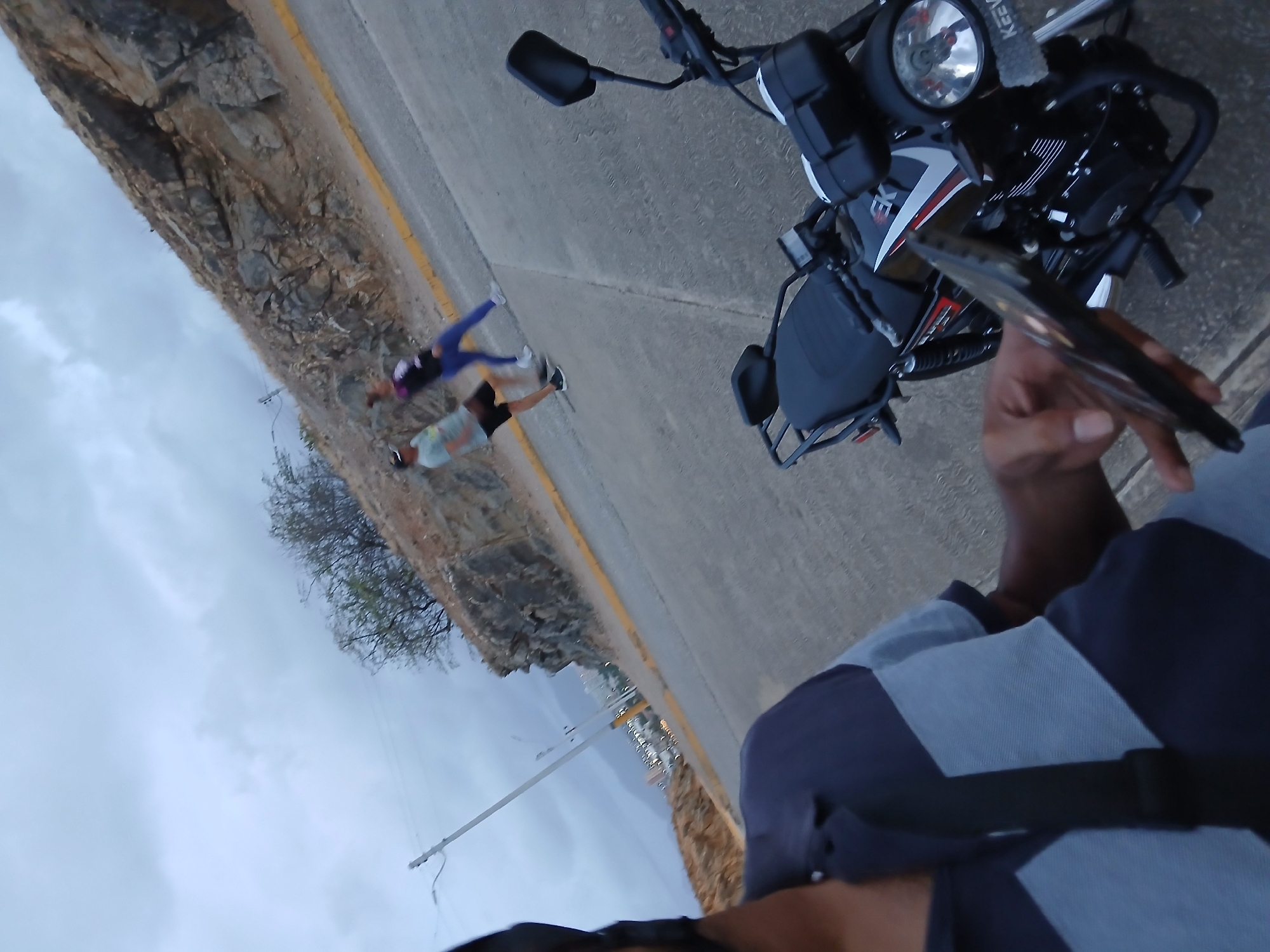 The image, taken from a first-person perspective, features a person standing next to a parked motorcycle on a road in what appears to be Juan Griego, Venezuela. Two individuals are running in the background, one wearing blue/purple activewear. A rock wall lines one side of the road, while the other side opens up to a view of the city and the sea. The sky is overcast. The person taking the photo is holding a phone in their hand.